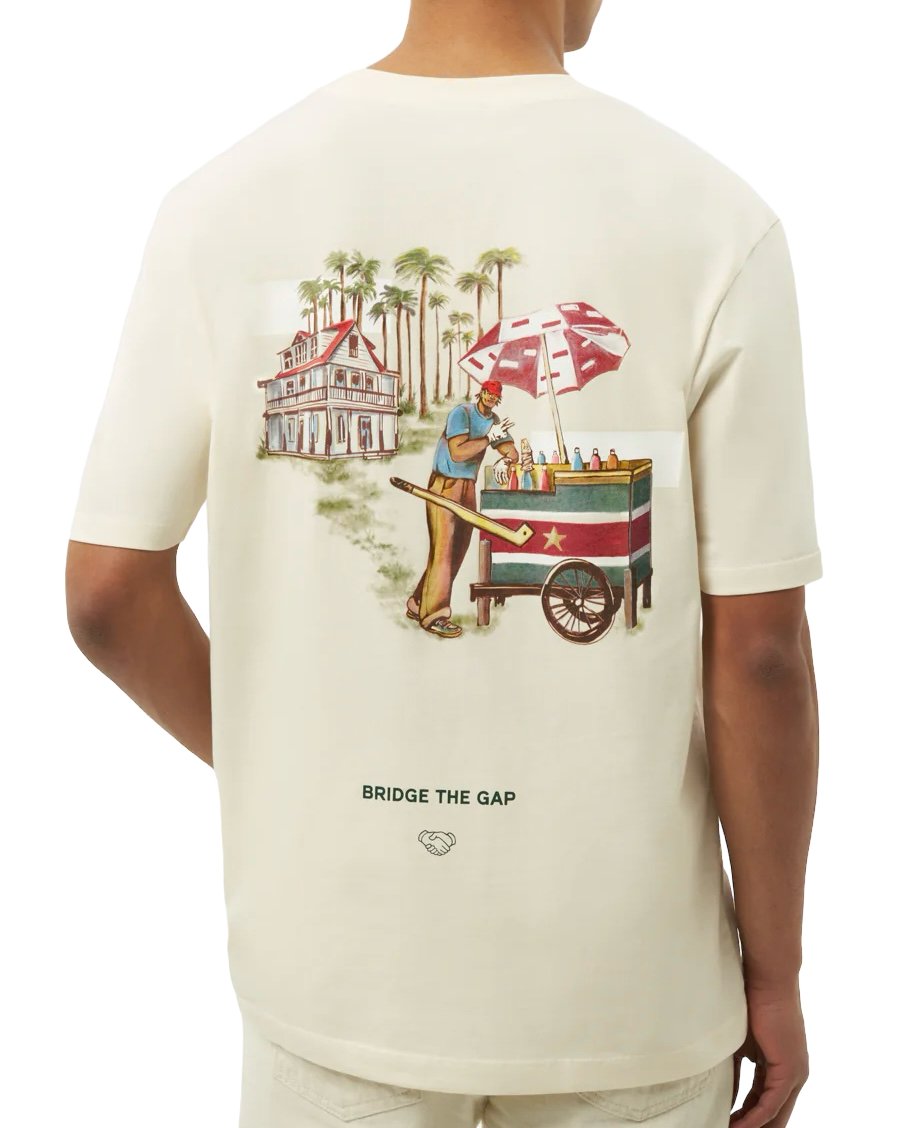Back view of a man wearing a FILLING PIECES ICE VENDOR ANTIQUE WHITE T-SHIRT with a colorful graphic of a person pushing a cart and a slogan "bridge the gap" printed below.