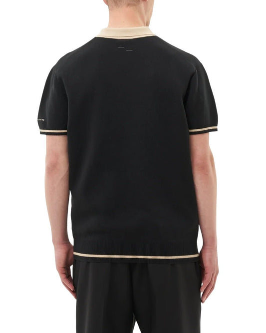 Rear view of a person wearing the FILLING PIECES KNIT POLO BLACK, a black short-sleeve knit polo with beige trim on the collar and sleeves, paired with black pants. The polo features FP branded buttons and is made from 100% cotton.