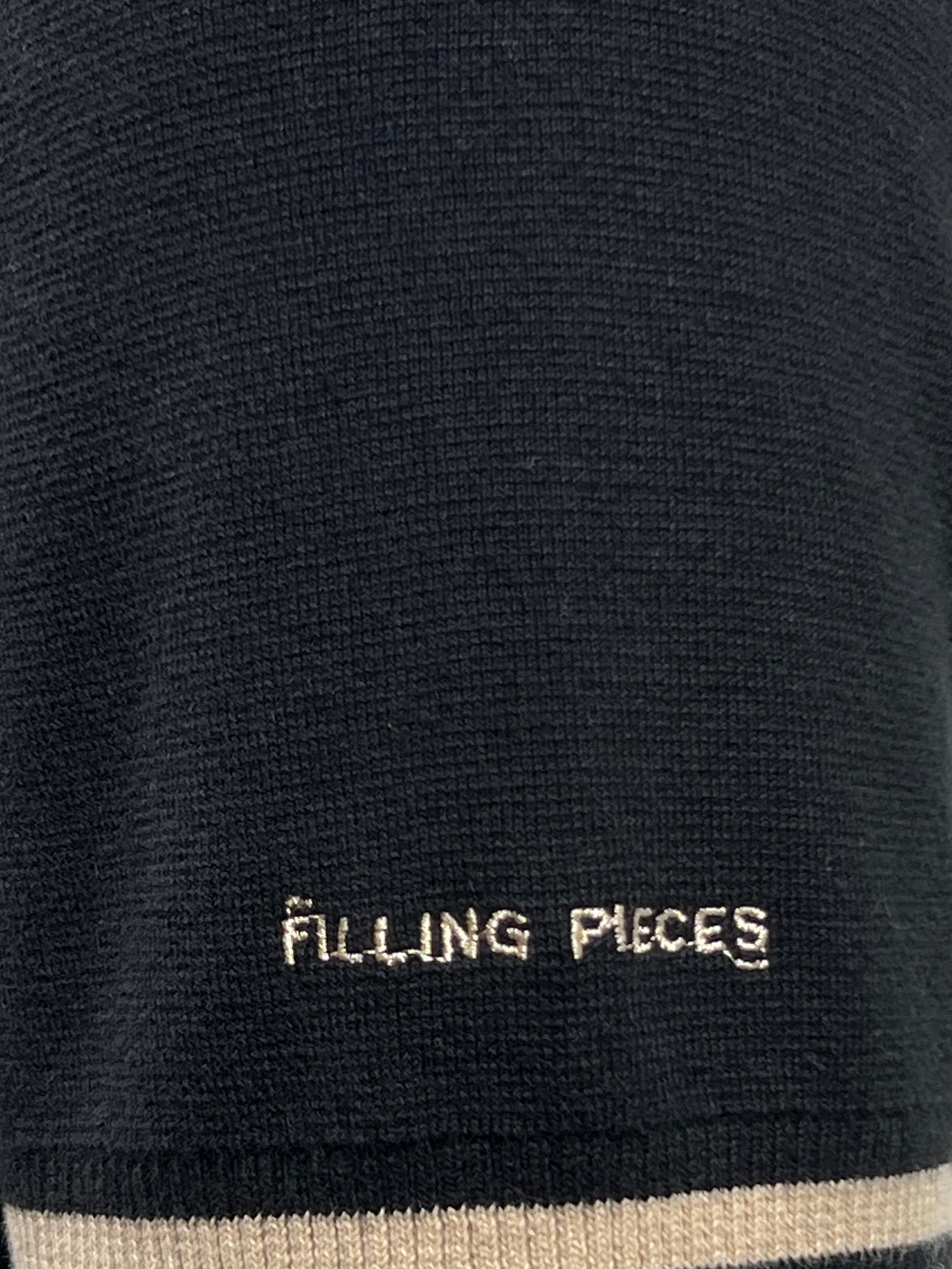 Close-up view of the FILLING PIECES KNIT POLO BLACK featuring black fabric with beige text that reads "FILLING PIECES" and a beige stripe at the bottom edge, reminiscent of the refined design found in 100% cotton knit garments from the brand FILLING PIECES.