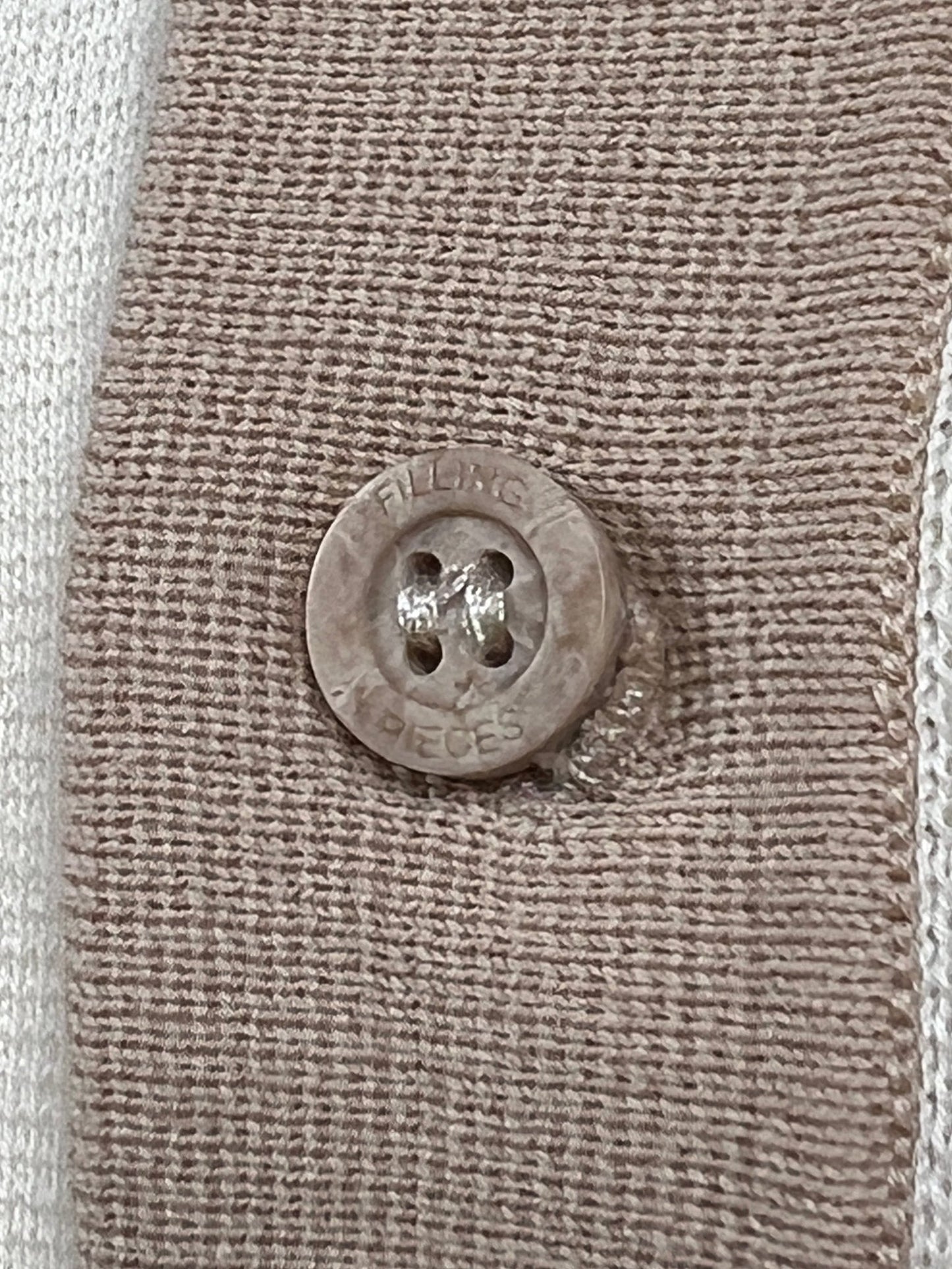 Close-up of a brown Filling Pieces branded button sewn onto a textured fabric strip of a Filling Pieces Knit Polo Rice Brown. The button has lettering indicating "FOUR HOLES 4 PIECES.