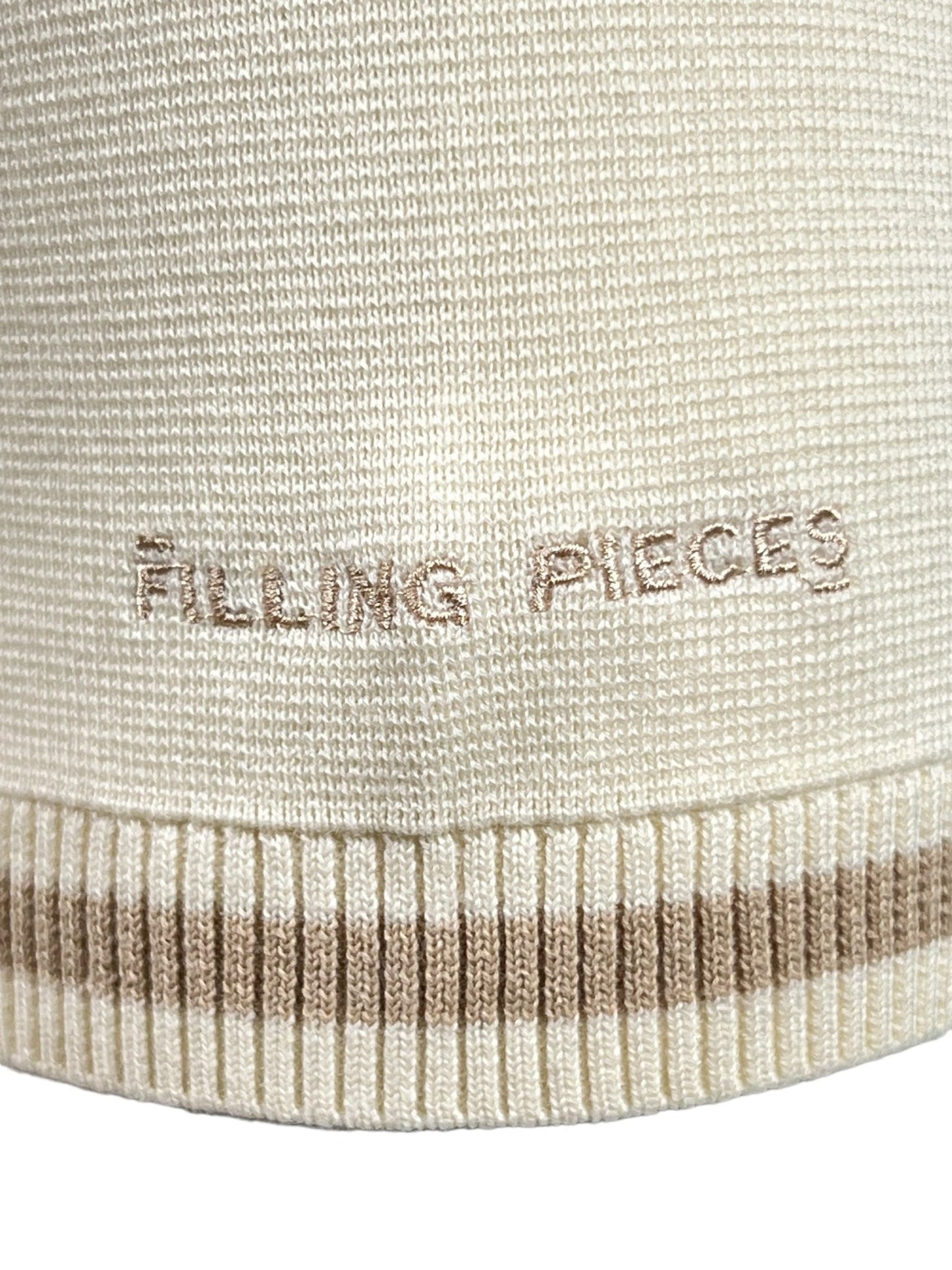 Close-up of the cream-colored knitted fabric of the FILLING PIECES KNIT POLO RICE BROWN, featuring 'FILLING PIECES' embroidered in beige. The bottom edge showcases a ribbed texture with brown and cream stripes, reflecting the meticulous craftsmanship typical of our knit polo collection.