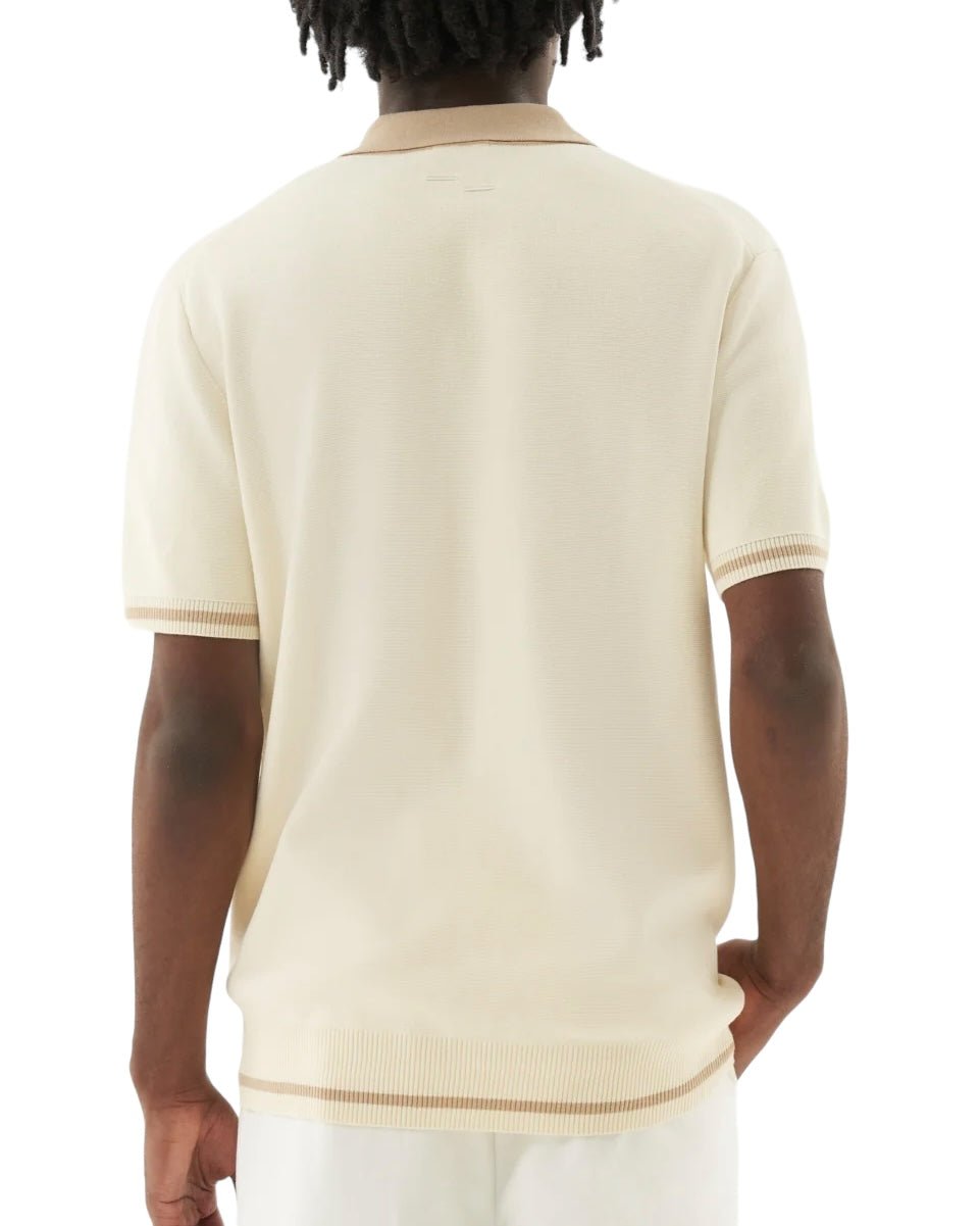 A person with short dreadlocks is seen from the back, wearing the FILLING PIECES KNIT POLO RICE BROWN shirt by FILLING PIECES. The cream short-sleeve knit polo is made of 100% cotton and features contrasting trim at the sleeves, collar, and hem. Notably, FP branded buttons add an extra touch of detail to the shirt.