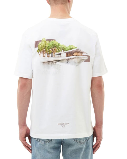 A person is wearing a FILLING PIECES PAVILION WHITE T-SHIRT, which features an illustration of a modern house surrounded by trees on the back. Seen from behind, the 100% organic cotton t-shirt from FILLING PIECES also sports a subtle FP chest logo and says "BRIDGE THE GAP" at the bottom.