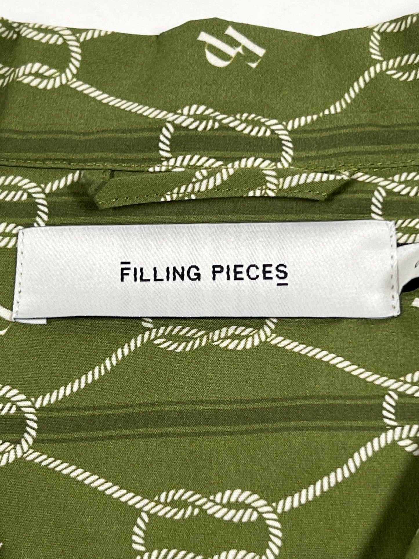 A close-up view of a clothing label with the text "FILLING PIECES" on an olive green patterned fabric of a FILLING PIECES RESORT MONOGRAM SHIRT OLIVE.