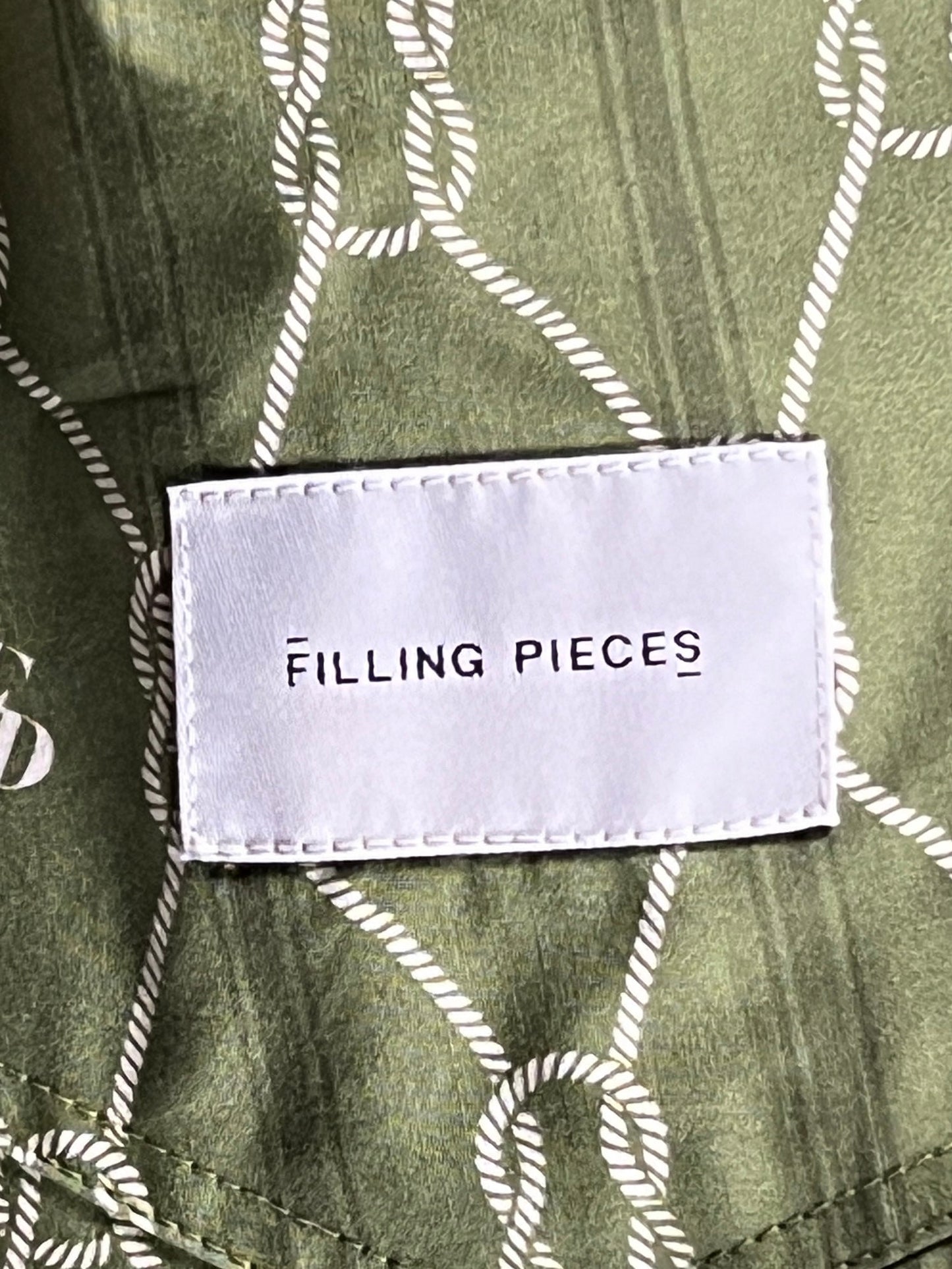 A clothing label with the text "FILLING PIECES" sewn onto FILLING PIECES RESORT MONOGRAM SHORT OLIVE shorts featuring an all-over print on 100% Organic Cotton fabric.
