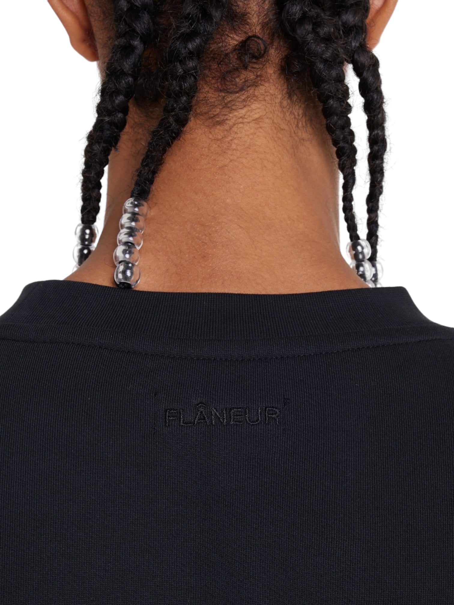 A close-up shows the back of a person's head wearing the FLANEUR Artist T-Shirt Black, a black cotton jersey with beaded braids and "FLÂNEUR" elegantly embroidered.
