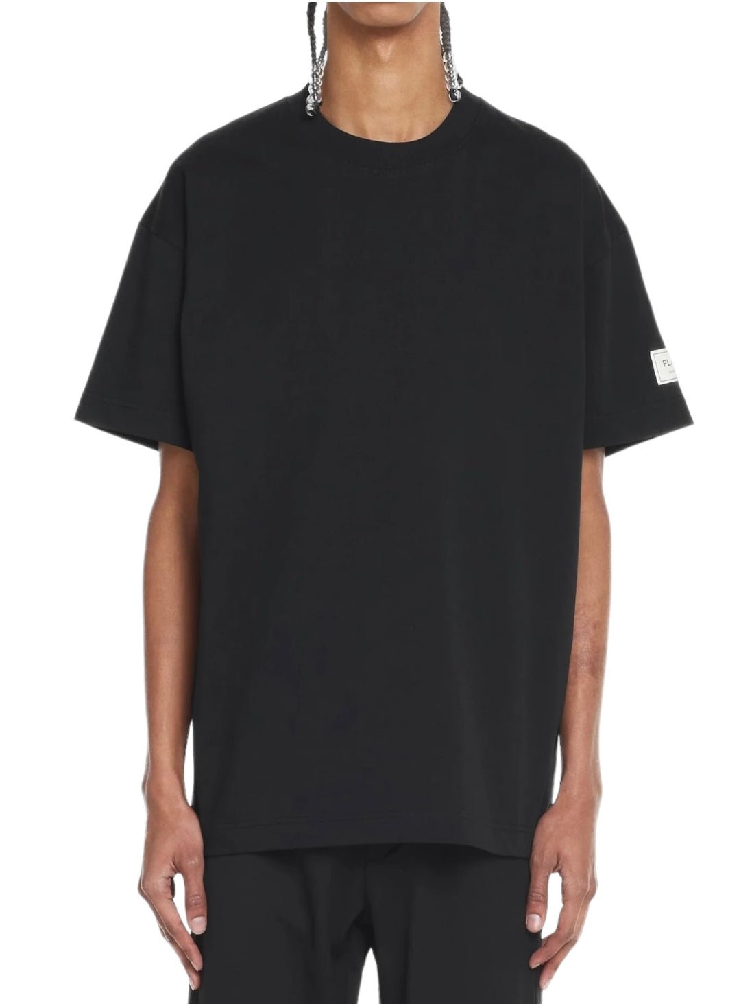 A person wearing a simple black FLANEUR ATELIER T-SHIRT BLACK crafted from soft cotton jersey, featuring short sleeves and a small white Flâneur-Atelier patch on the left sleeve. The individual has braided hair.