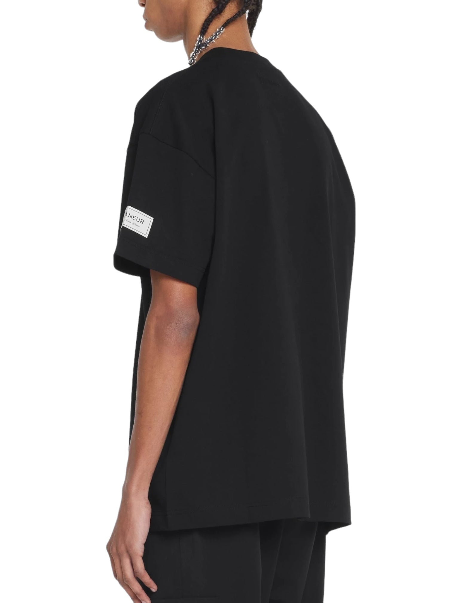 Wearing the Flaneur Atelier T-Shirt Black, a person stands sideways in a soft cotton jersey. The FLANEUR tee features a subtle sleeve patch.