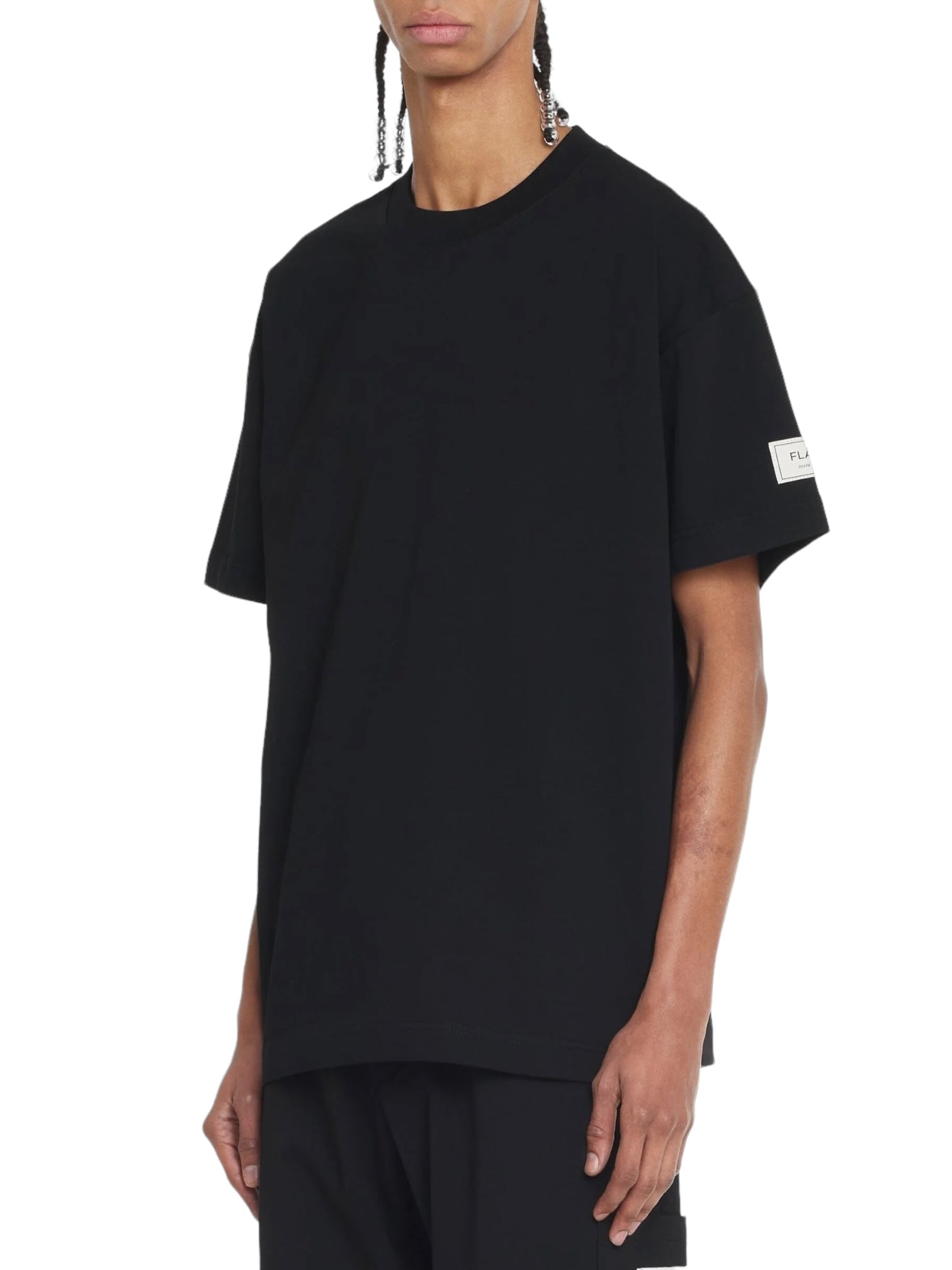 The individual is wearing a Flaneur Atelier T-Shirt in black from FLANEUR, made of soft cotton jersey, featuring a small patch on the sleeve.