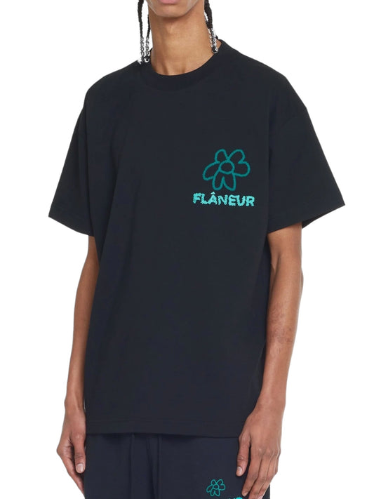 Person wearing the FLANEUR FLOWER DOODLE T-SHIRT BLACK with a green flower doodle graphic and the word "FLÂNEUR" in Flâneur graphic lettering on the chest, paired with black pants featuring a small green flower logo.