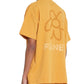 Person wearing an oversized mustard-colored "Flaneur Flower Doodle T-Shirt Org" by FLANEUR, featuring a flower doodle graphic and the word "FUNE" on the back.