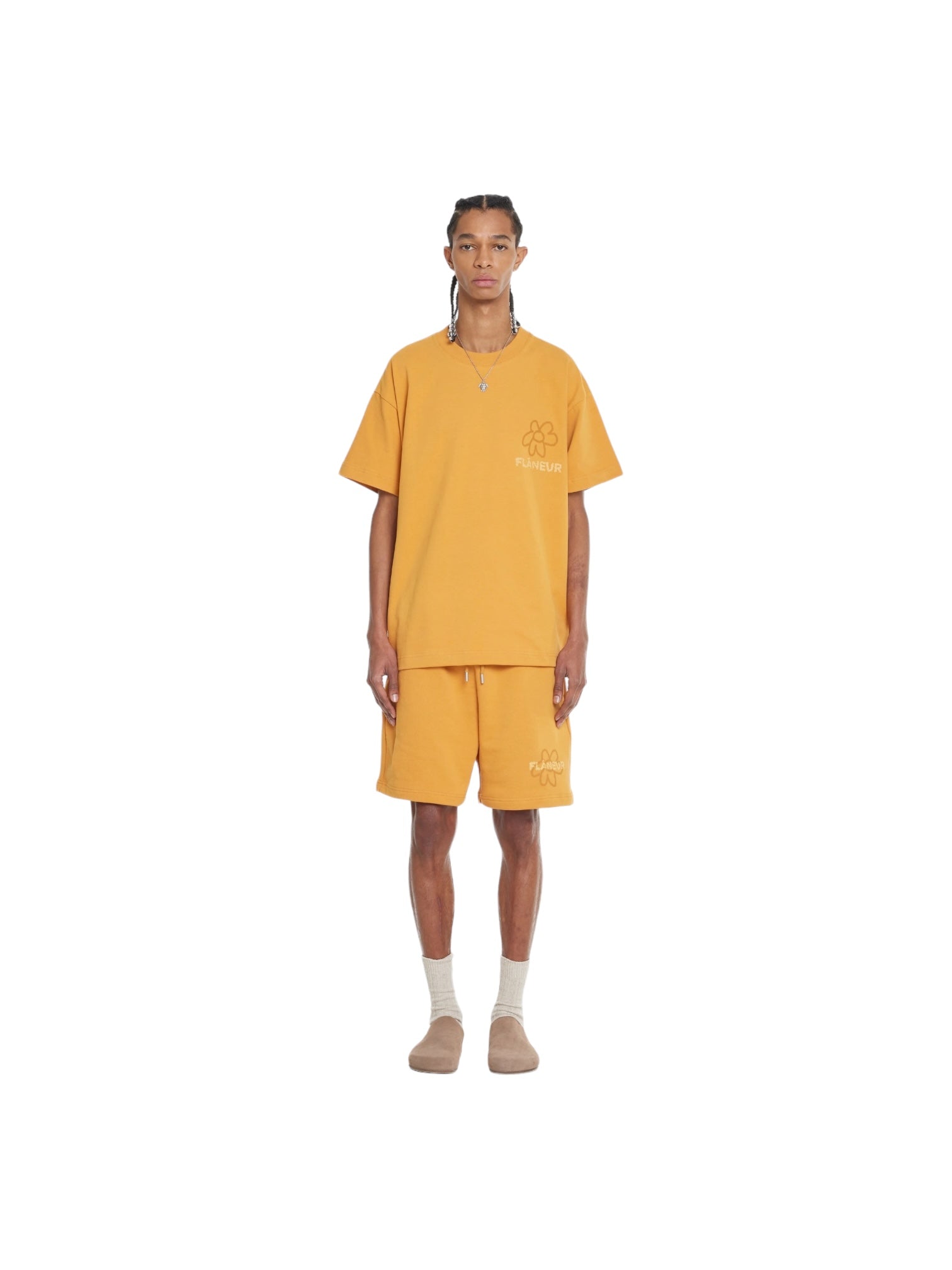 A person stands confidently against a pristine white background, wearing a vibrant orange Flaneur Flower Doodle T-Shirt by FLANEUR, adorned with a playful flower doodle graphic. They have paired it with matching cotton jersey bottoms, white socks, and beige slippers.