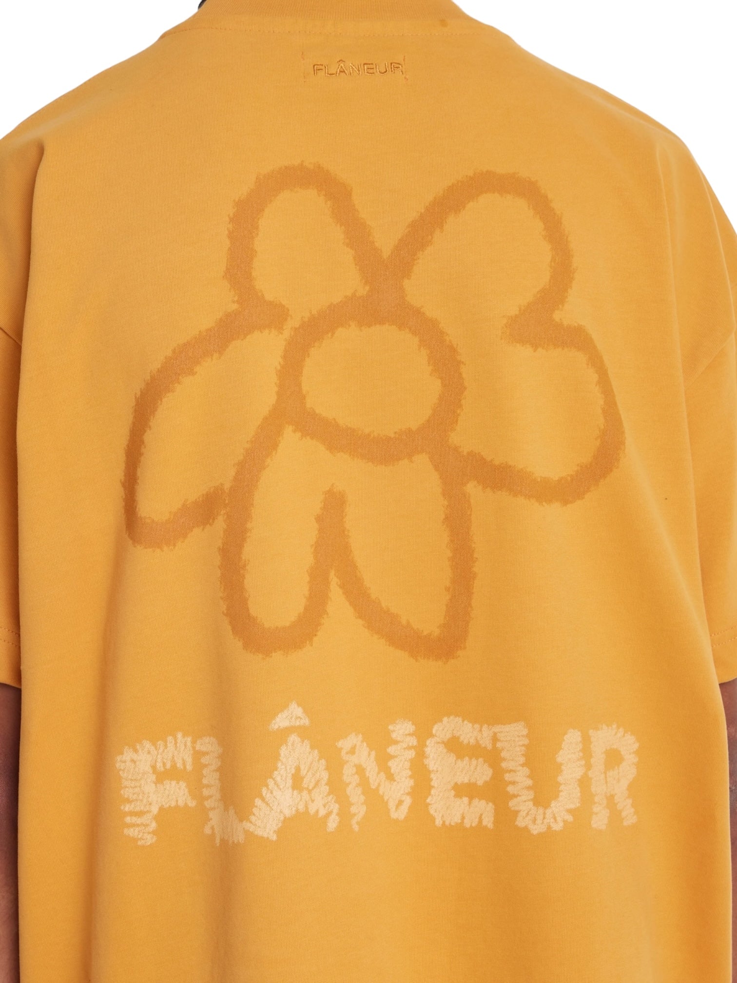 A person wearing the Flaneur Flower Doodle T-Shirt Org in yellow, featuring a large flower doodle graphic and the brand name "FLANEUR" on the back.