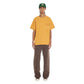 A person wearing the FLANEUR Flaneur Flower Doodle T-Shirt Org in yellow, along with brown pants and a green cap, stands against a plain white background.