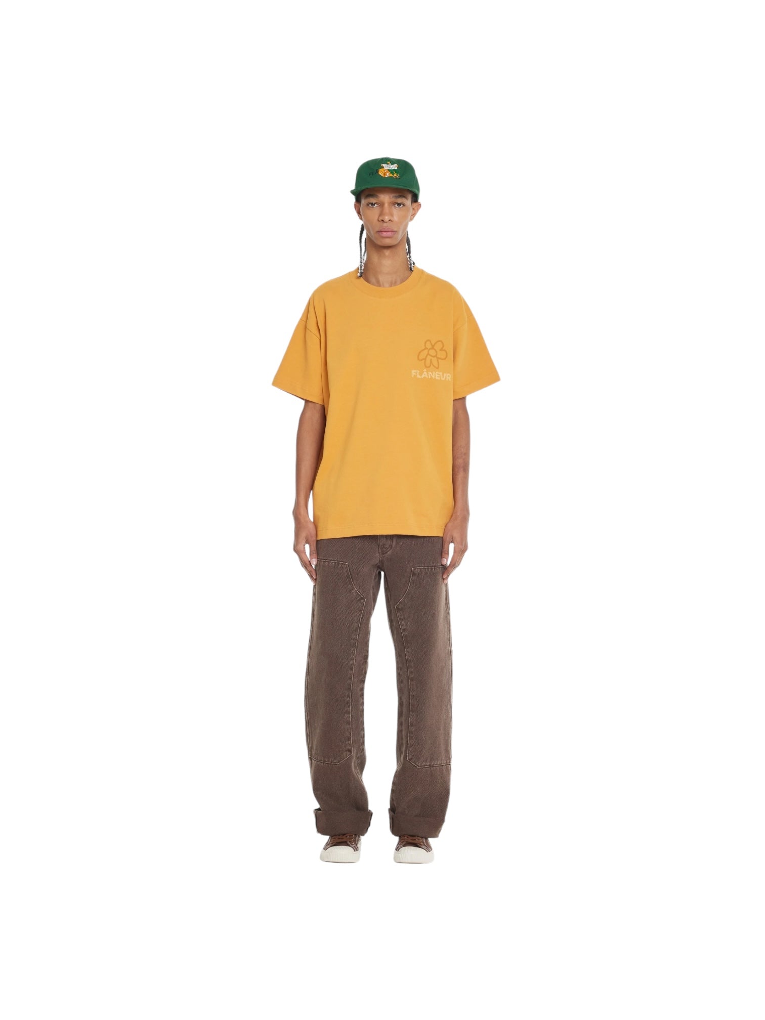 A person wearing the FLANEUR Flaneur Flower Doodle T-Shirt Org in yellow, along with brown pants and a green cap, stands against a plain white background.