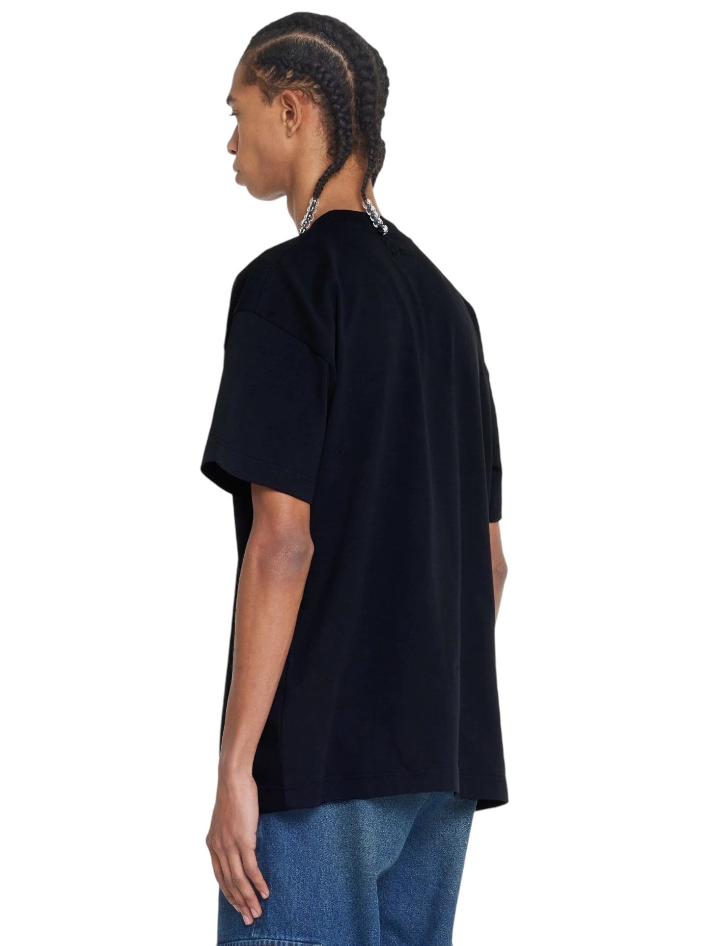 Person with braided hair wearing a black FLANEUR FLOWER STALK T-SHIRT made from cotton jersey and blue jeans, viewed from the side with their back slightly turned towards the camera.