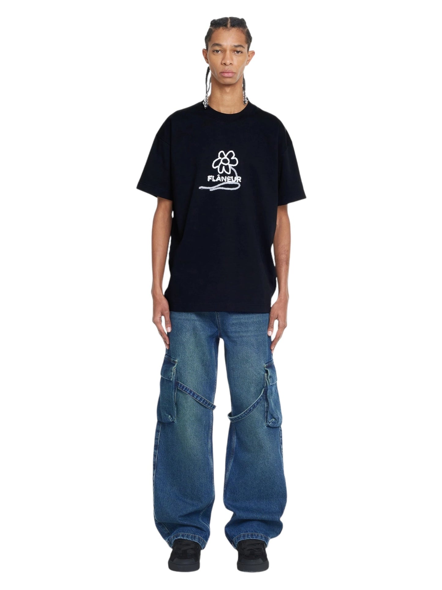 A person stands facing forward wearing a black Flaneur Flower Stalk T-Shirt made of soft cotton jersey, paired with blue baggy jeans featuring zipper details. The individual has long braided hair and black shoes.