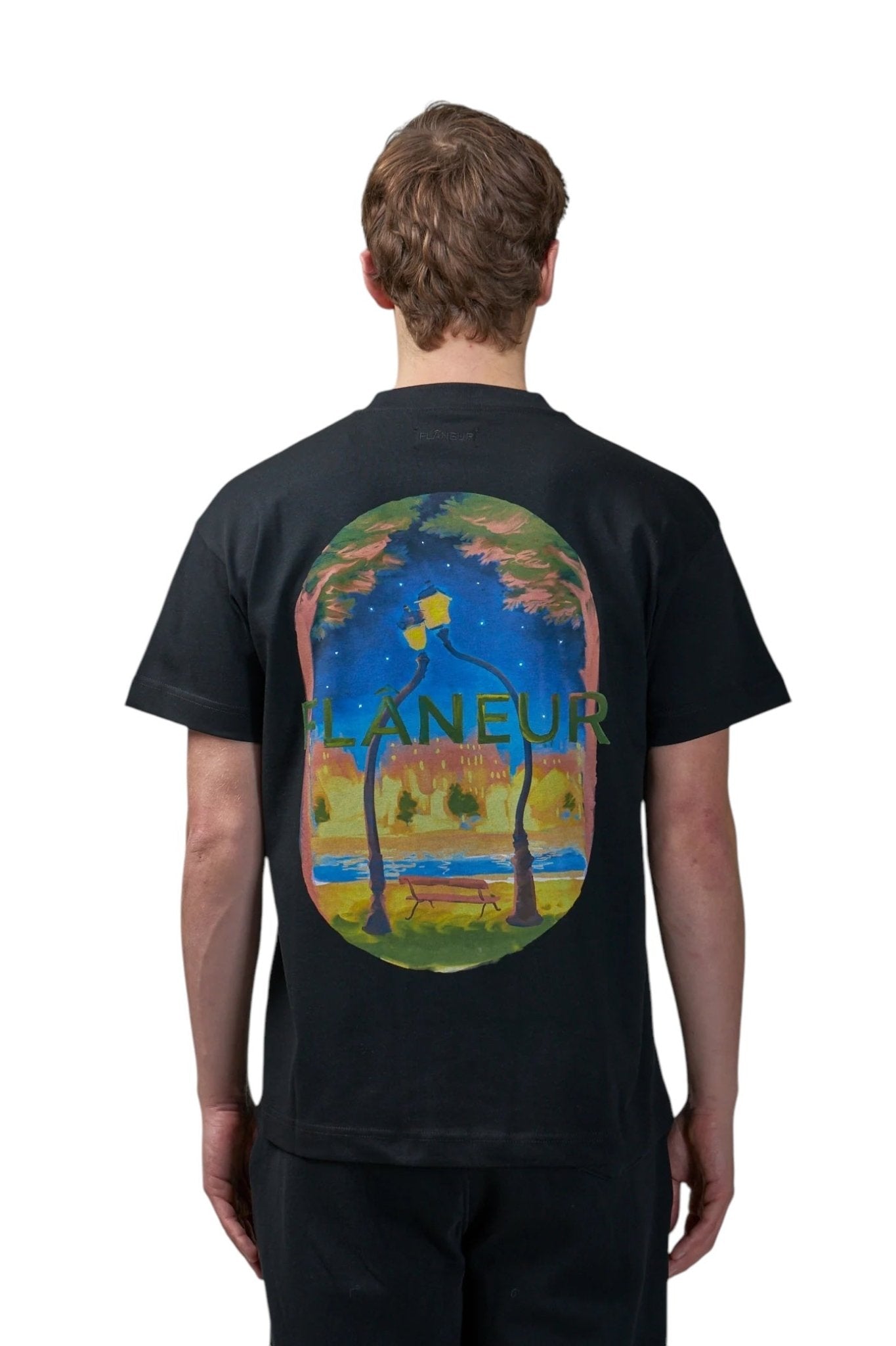 Someone wears the FLANEUR Flaneur Lanterns of Love T-Shirt Black, featuring a colorful night scene with trees, a lamppost, love lanterns, a river, and "L'Aneur" printed on the back.