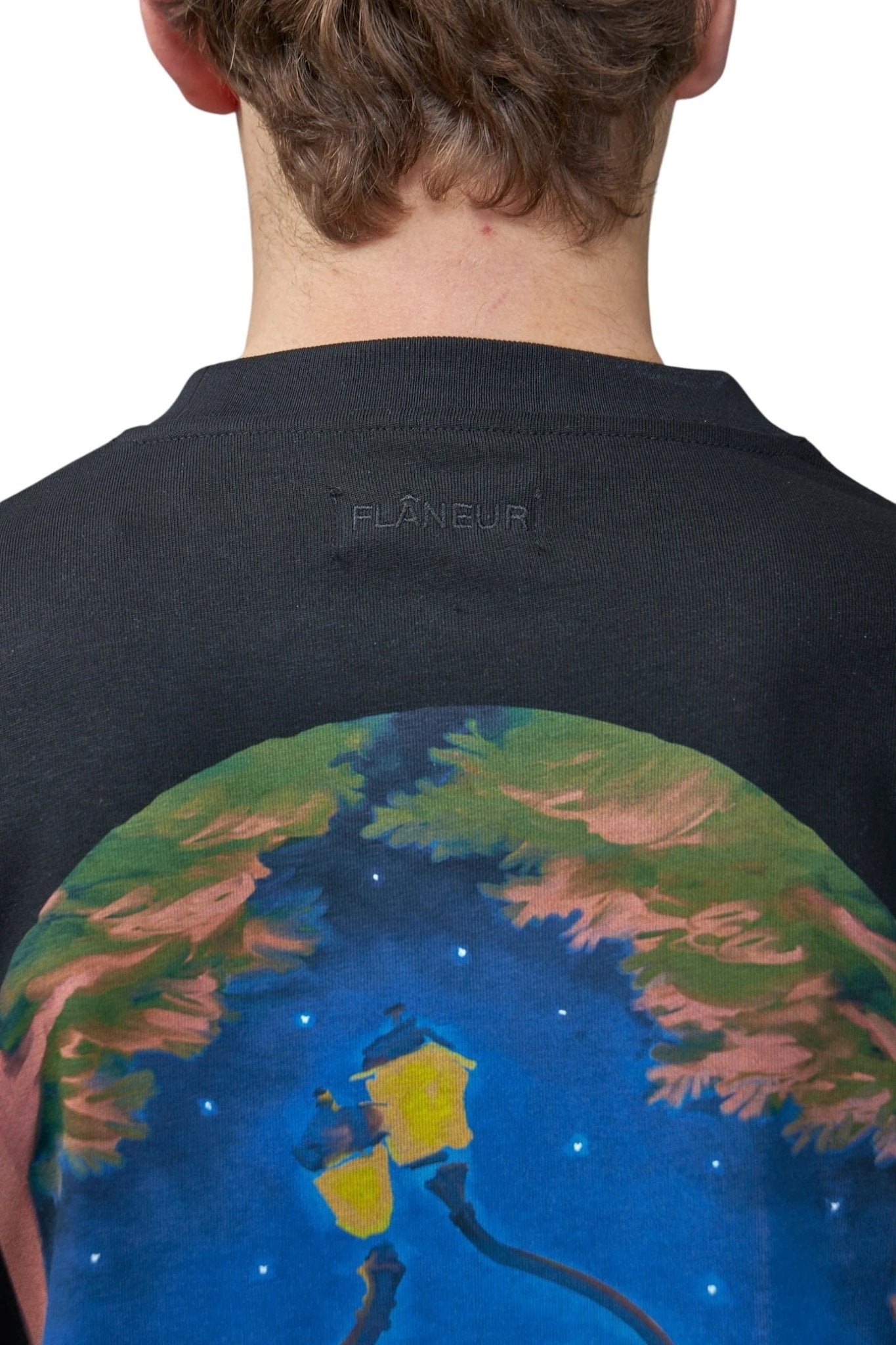 A person wears the FLANEUR Lanterns of Love T-Shirt Black, featuring a vibrant streetlamp and tree design on the back with "FLÂNEUR" embossed below the collar.