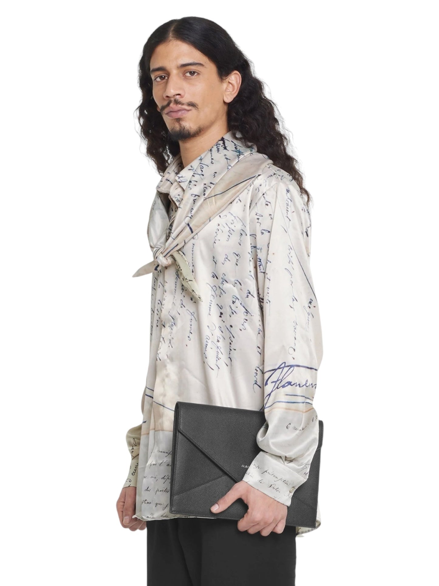 A man with long hair and a beard, wearing a beige FLANEUR LETTER VISCOSE SHIRT by FLANEUR featuring a script pattern, holds a black envelope-style clutch and stands against a plain white background.