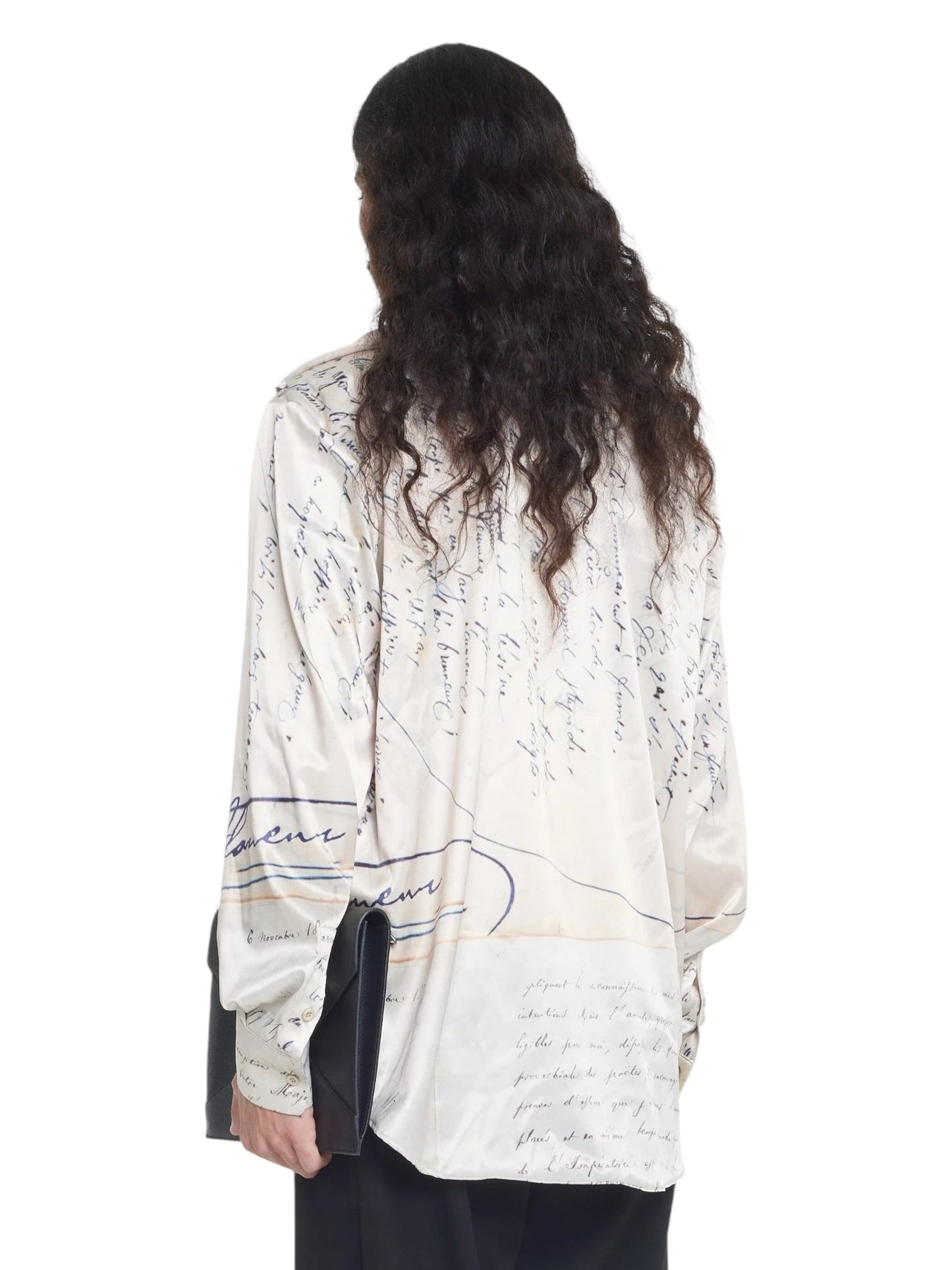 Person with long, wavy hair wearing the FLANEUR LETTER VISCOSE SHIRT BEIGE, featuring handwritten text patterns, viewed from the back.