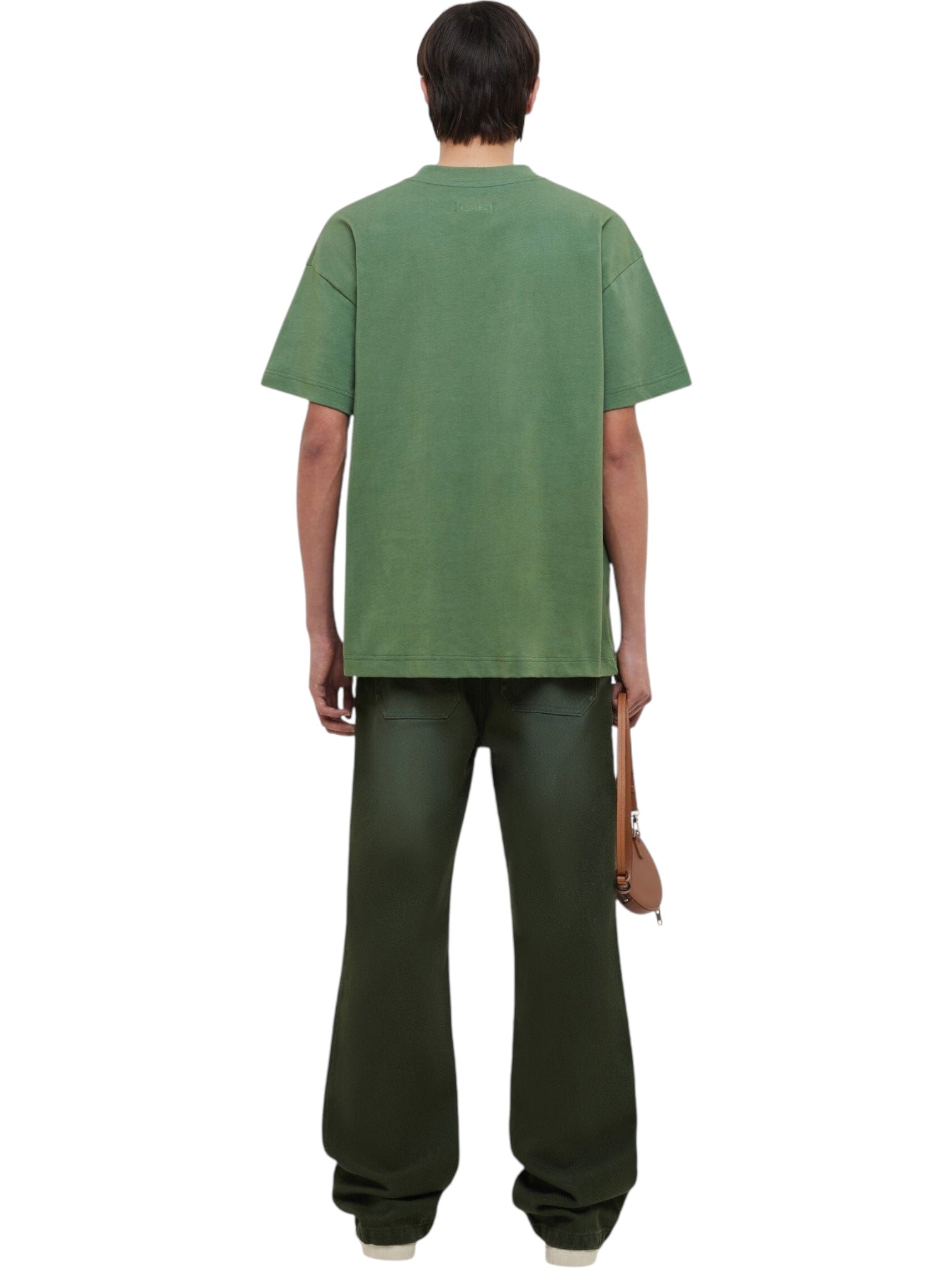 A person stands facing away, wearing a FLANEUR Flaneur Logo T-Shirt Khaki with a front print, wide dark green pants, and holding a small brown bag in their right hand.