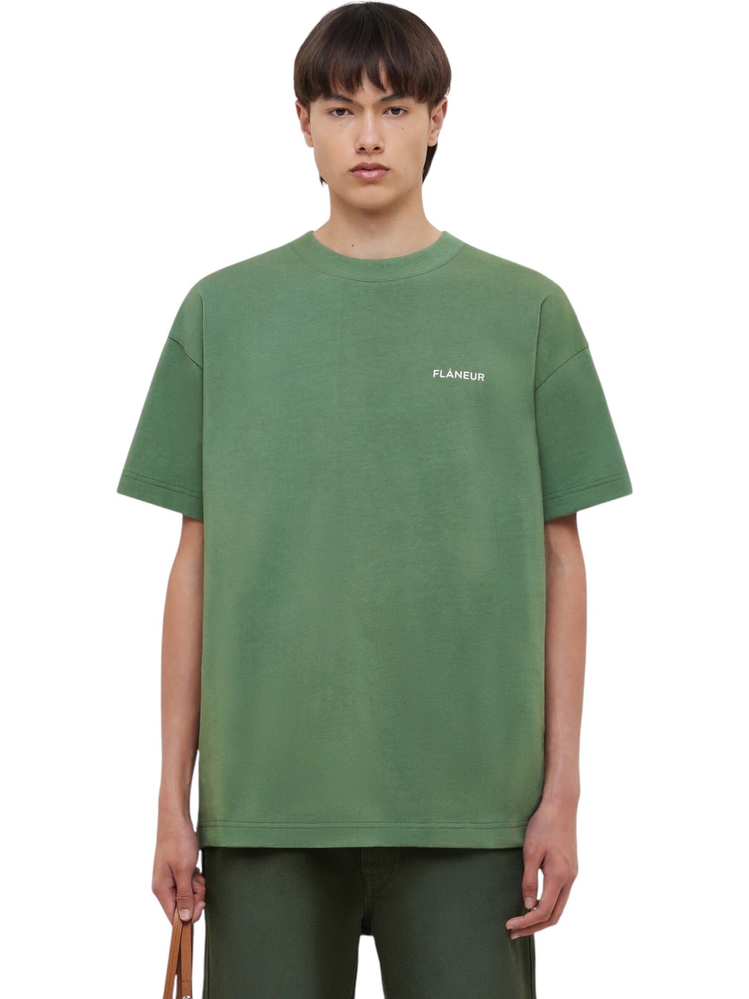 A person wears a loose-fitting FLANEUR Flaneur Logo T-Shirt Khaki with "FLÂNEUR" white print on the left chest and green pants, standing against a plain background.