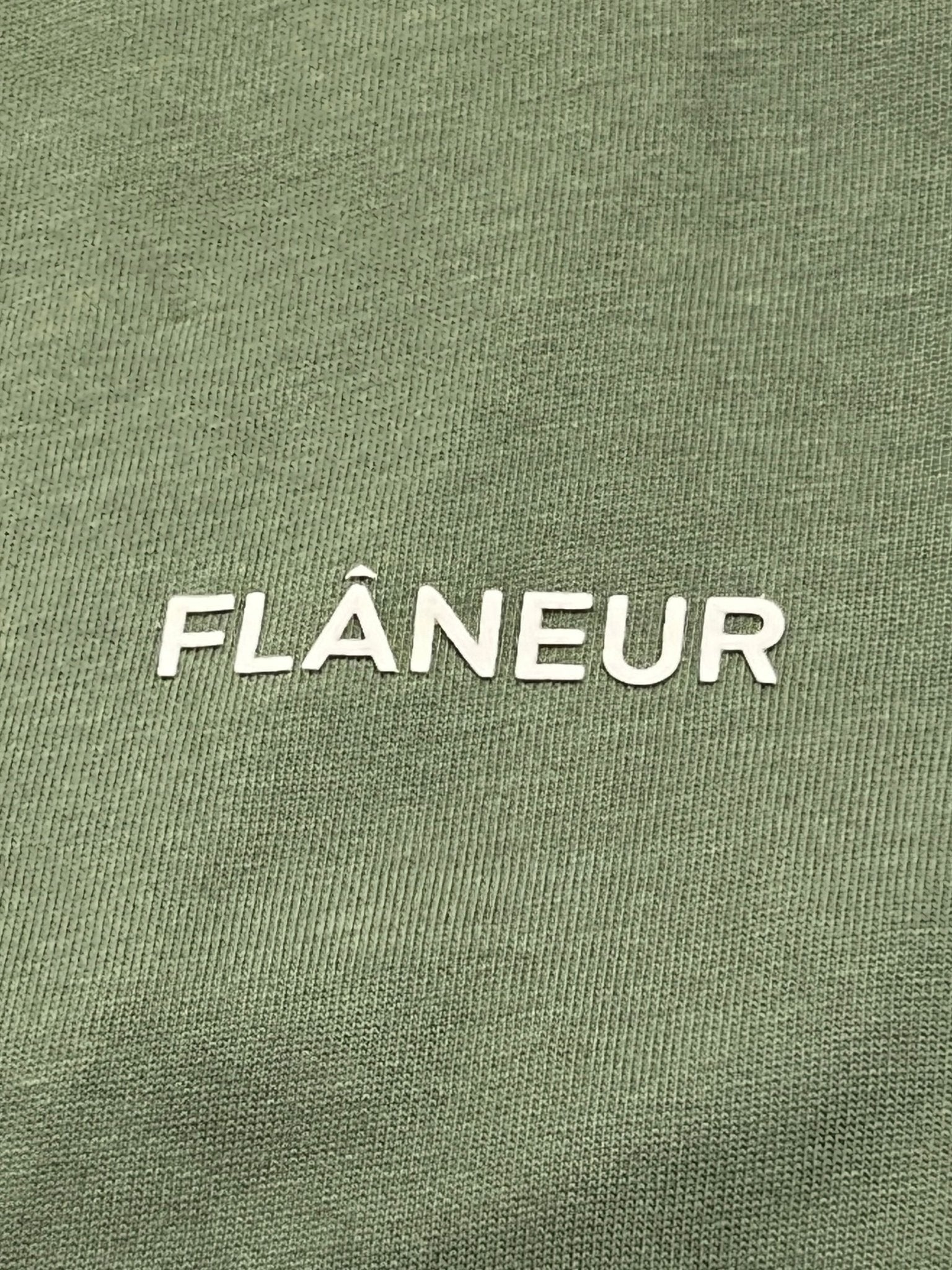 The Flaneur Logo T-Shirt Khaki by FLANEUR features "FLÂNEUR" in bold white caps on textured khaki cotton for a stylish, modern look.