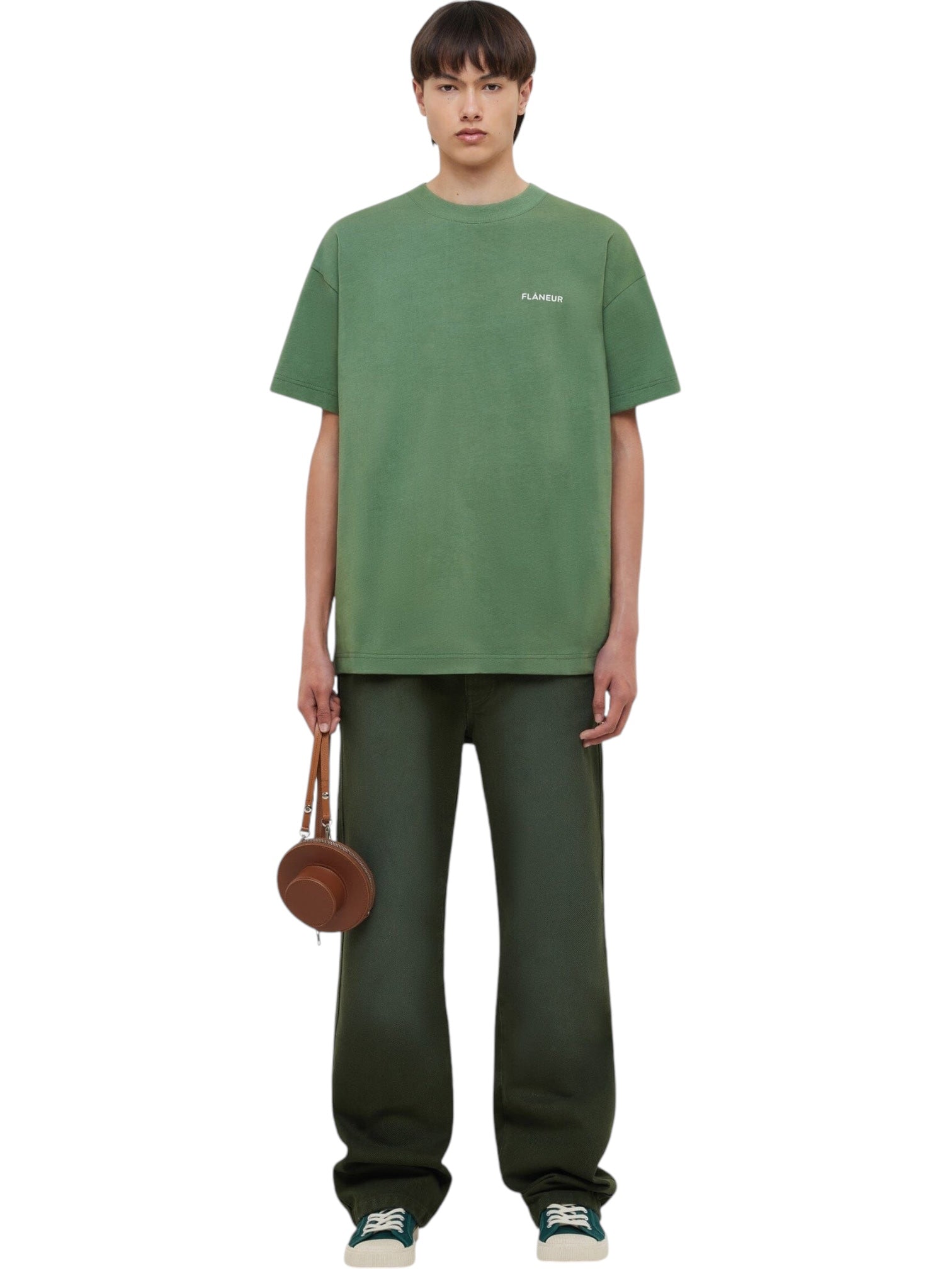 A person stands facing forward, wearing a FLANEUR Flaneur Logo T-Shirt Khaki with a front print, dark green pants, green sneakers, and holding a small round brown bag in their left hand.