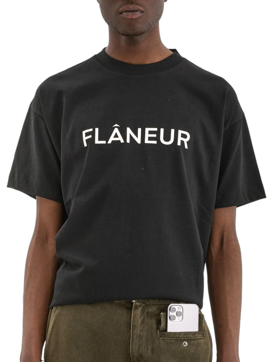 A person wearing a FLANEUR Printed Logo T-Shirt in black with "FLÂNEUR" in white text has a smartphone partially tucked into the front pocket of their khaki pants.