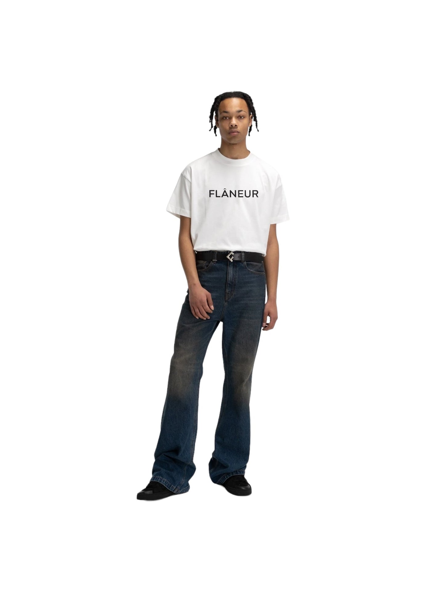 A person stands against a plain background, wearing the Flaneur Printed Logo T-Shirt in white, featuring "FLÂNEUR" across the front, paired with blue jeans.