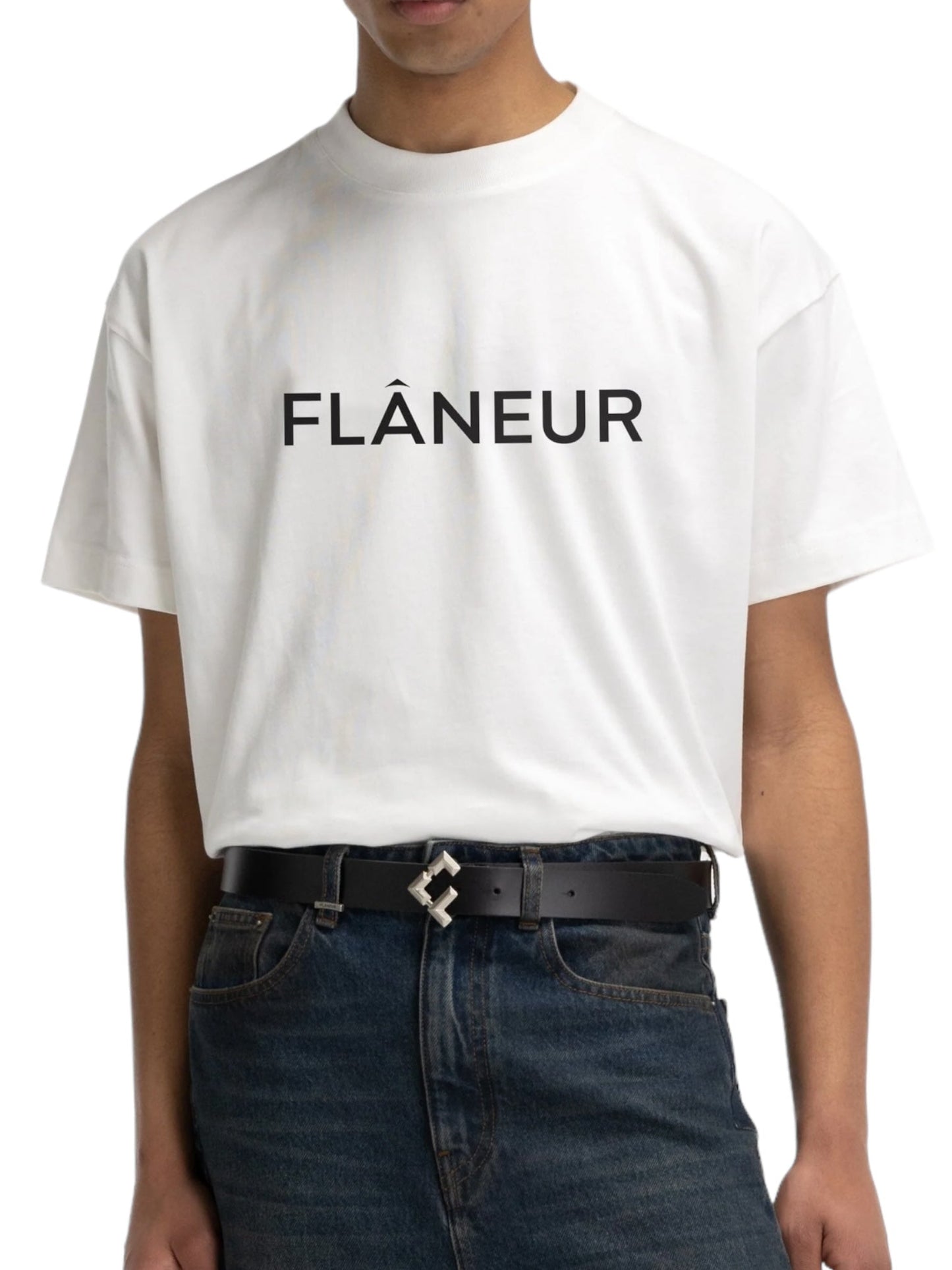 A person wears the Flaneur Printed Logo T-Shirt in white, embossed with "FLÂNEUR" lettering, tastefully paired with jeans and a black belt.