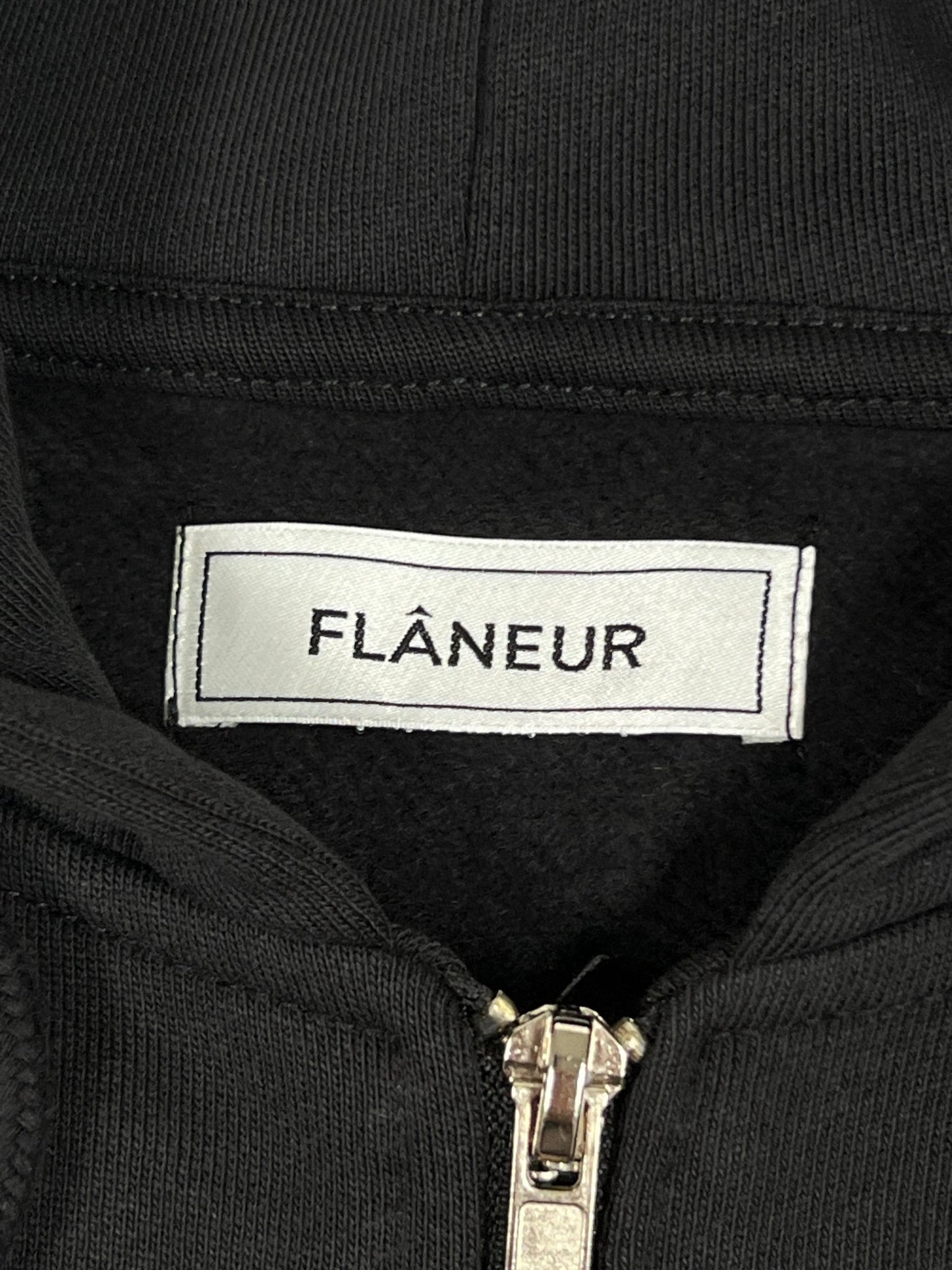 Close-up image of a clothing label with the word "FLÂNEUR" on a black fabric, positioned above an unzipped metallic zipper. This stylish detail is featured on the FLANEUR SIGNATURE ZIP-UP HOODIE BLACK, made from 100% cotton and perfect for casual elegance.