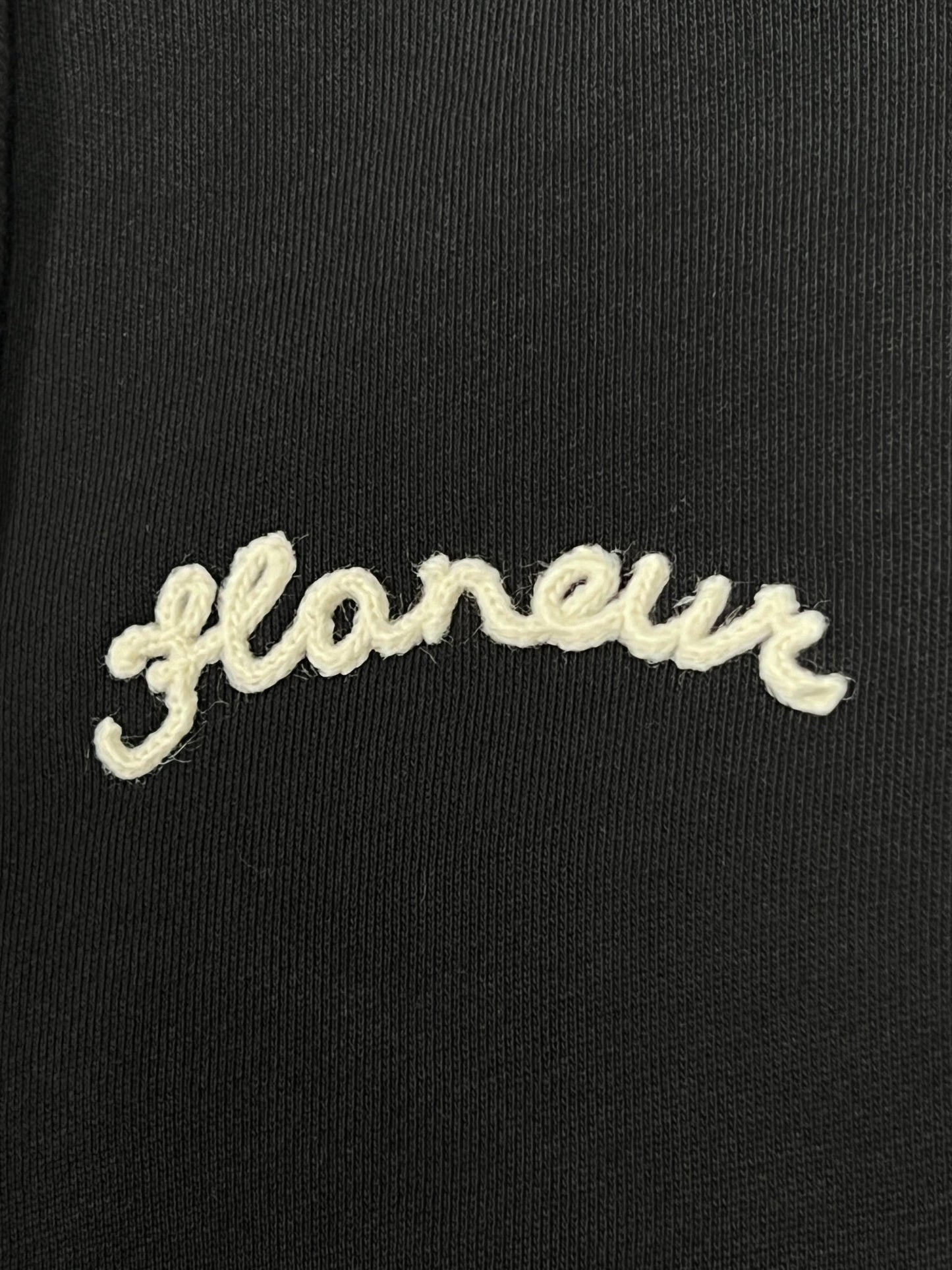 Close-up of the word "Flaneur" embroidered in white cursive on black fabric, part of the FLANEUR SIGNATURE ZIP-UP HOODIE BLACK made from 100% cotton.