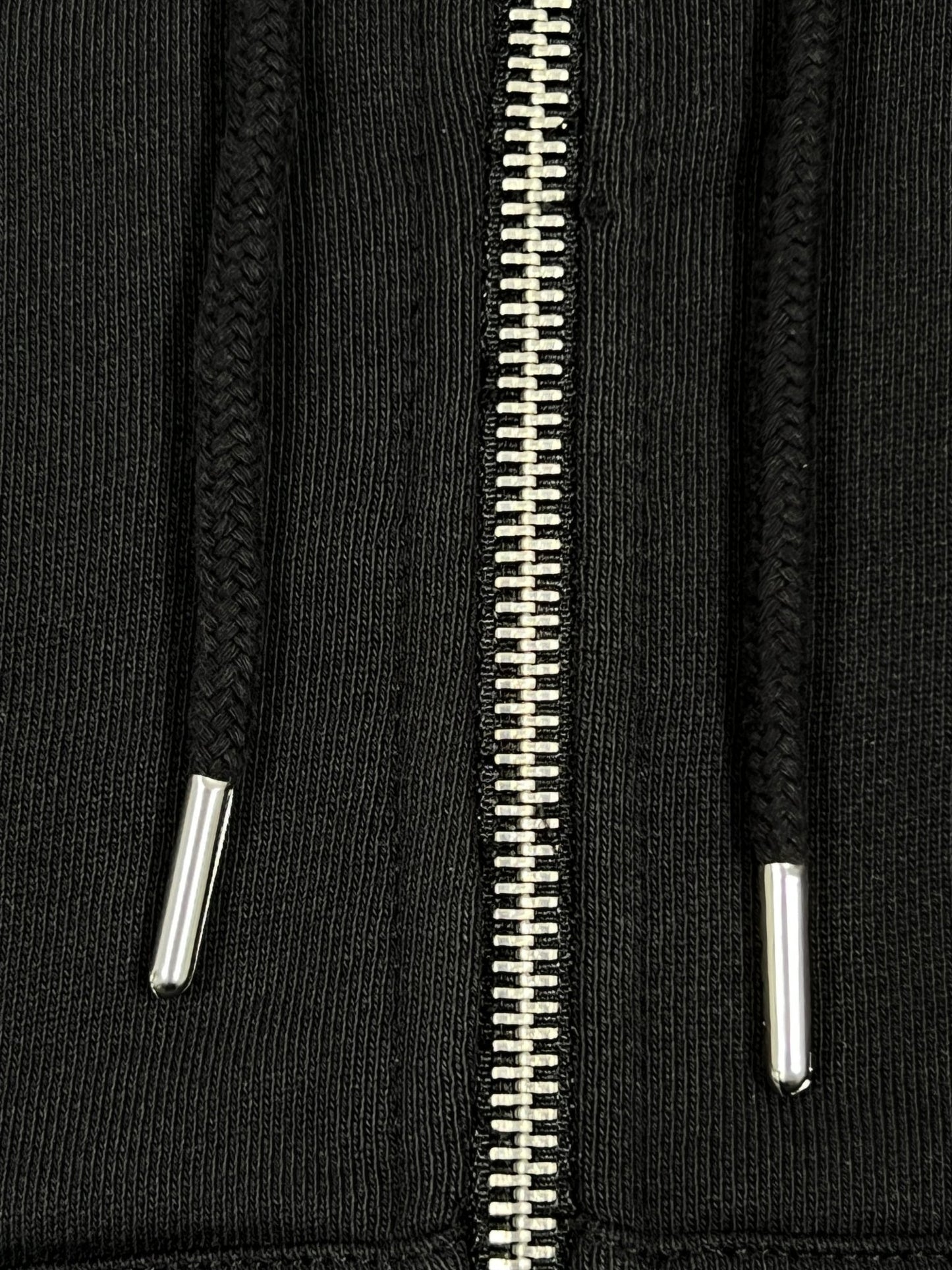 Close-up of the black zipper and drawstrings on the FLANEUR SIGNATURE ZIP-UP HOODIE BLACK, made from 100% cotton. The fabric appears ribbed, with clearly visible metallic zipper teeth and drawstring tips.