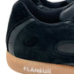 Close-up of the FLANEUR TIVOLI RUNNER BLACK, featuring black suede with matching black laces and a light brown rubber sole. The sneaker prominently displays the white inscription "FLANEUR" near the heel, combining an exquisite blend of style and eco-conscious design through sustainably sourced materials.