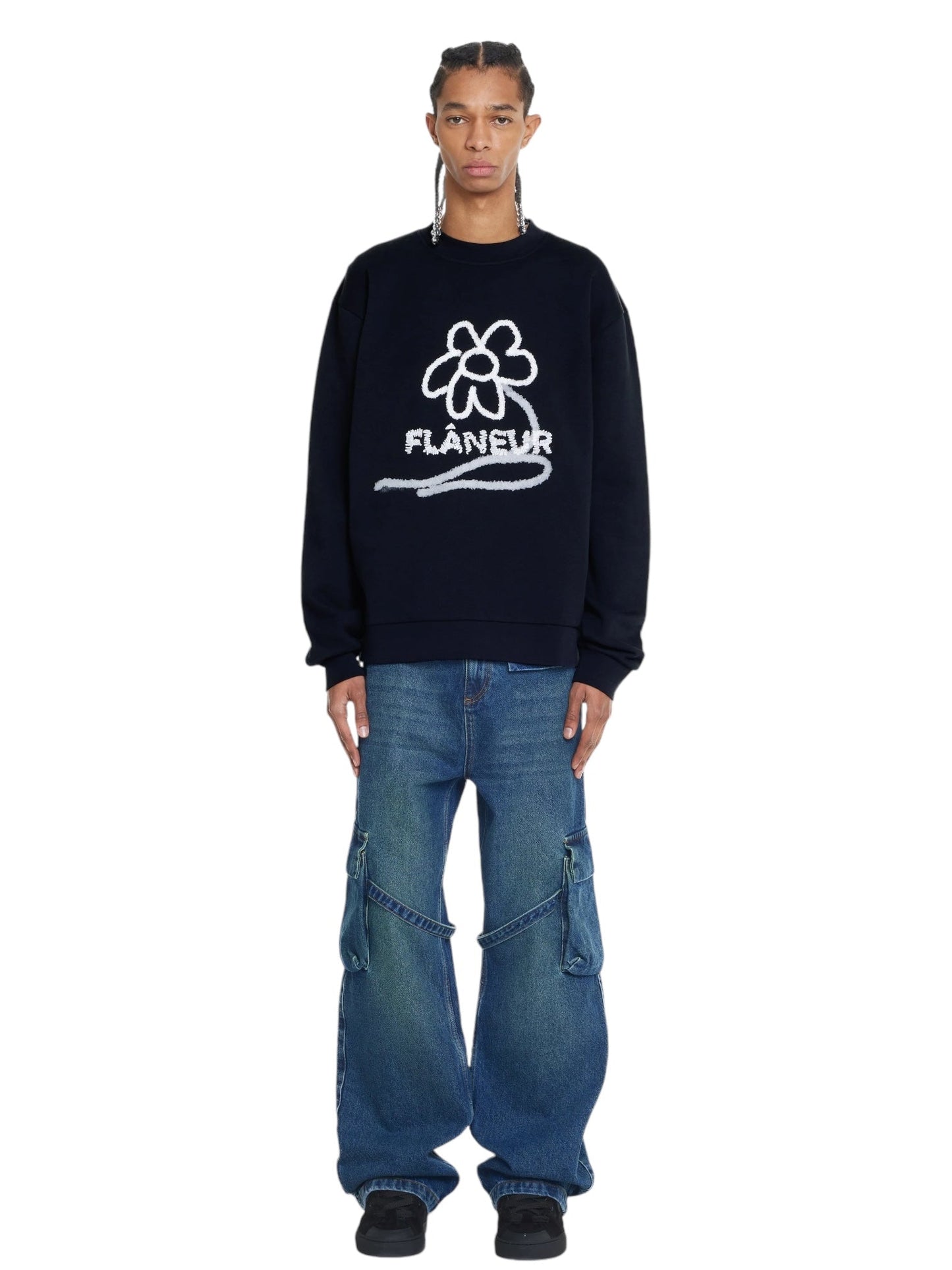 A person stands against a plain white background, wearing the FLANEUR FLOWER STALK SWEATER in black, made of soft cotton jersey and adorned with a white floral doodle graphic along with the word "Flâneur," paired with blue baggy jeans and black shoes.