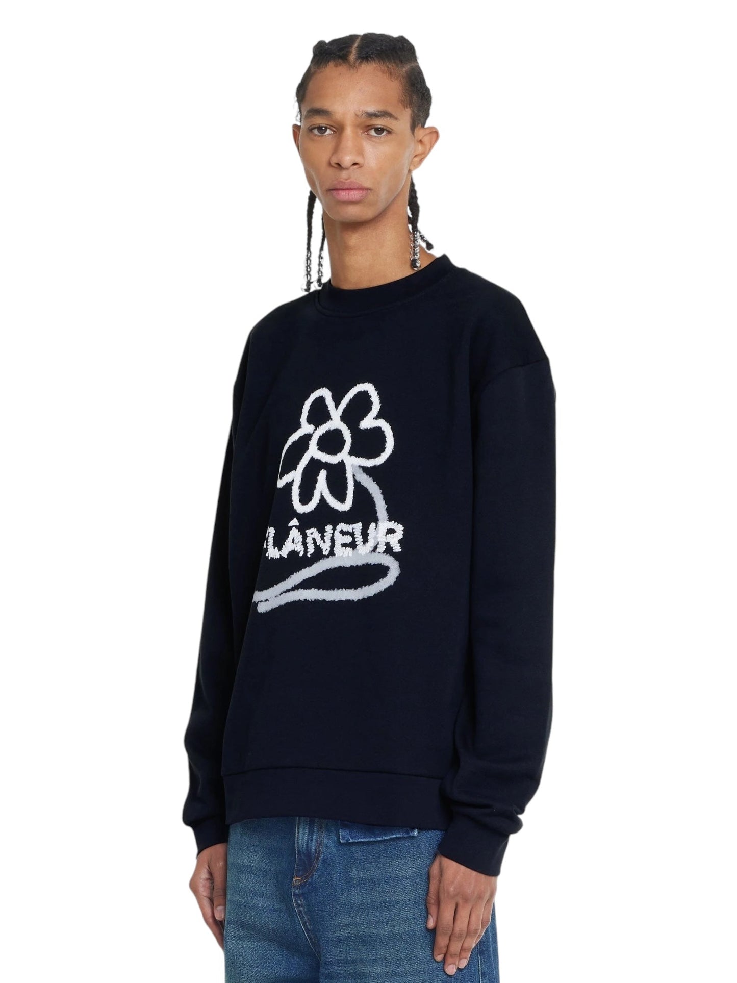 A person with braided hair is wearing the FLOWER STALK SWEATER BLACK by FLANEUR, crafted from soft cotton jersey, featuring a white flower graphic and the word "FLÂNEUR" across the front, paired with blue jeans.