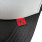Close-up of the FREE REFILLS Free Refills Harley Trucker (FR-C3-39) hat in white/black, featuring a small red tag with black graphic symbols.