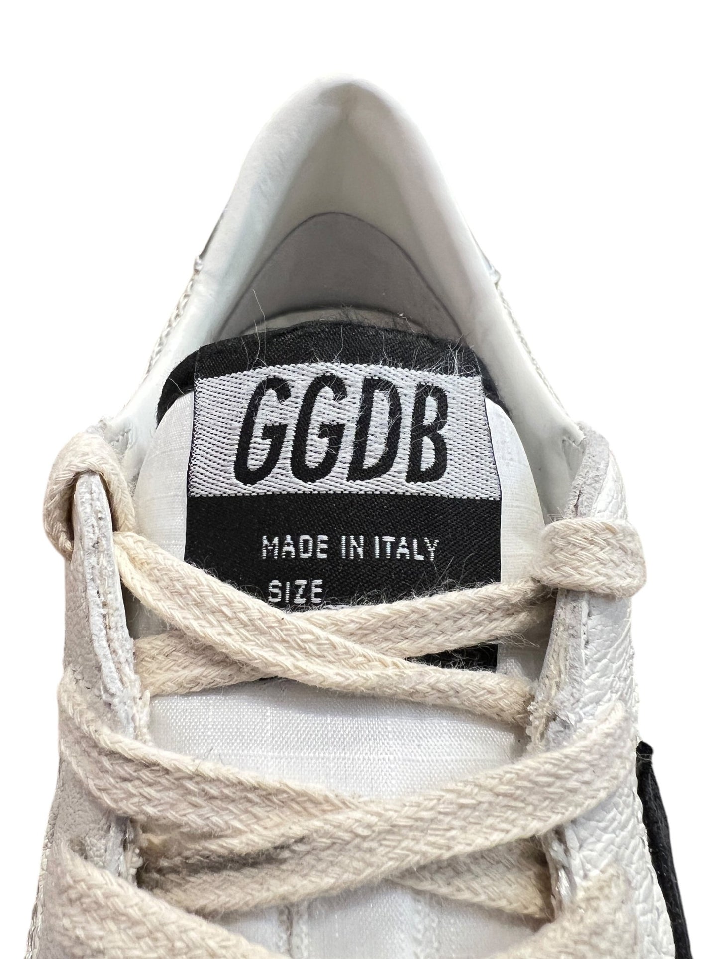 Close-up of a Golden Goose GMF00117 Ball Star sneaker tongue displaying "GGDB" and "Made in Italy." Partially visible white laces complement the design, with hints of a rubber sole peeking through.