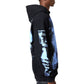 A young man stands in profile wearing the GUAPI Guapi Black Revived Hoodie V2 with blue graphic prints and dark patterned pants, showcasing modern streetwear style against a white background.