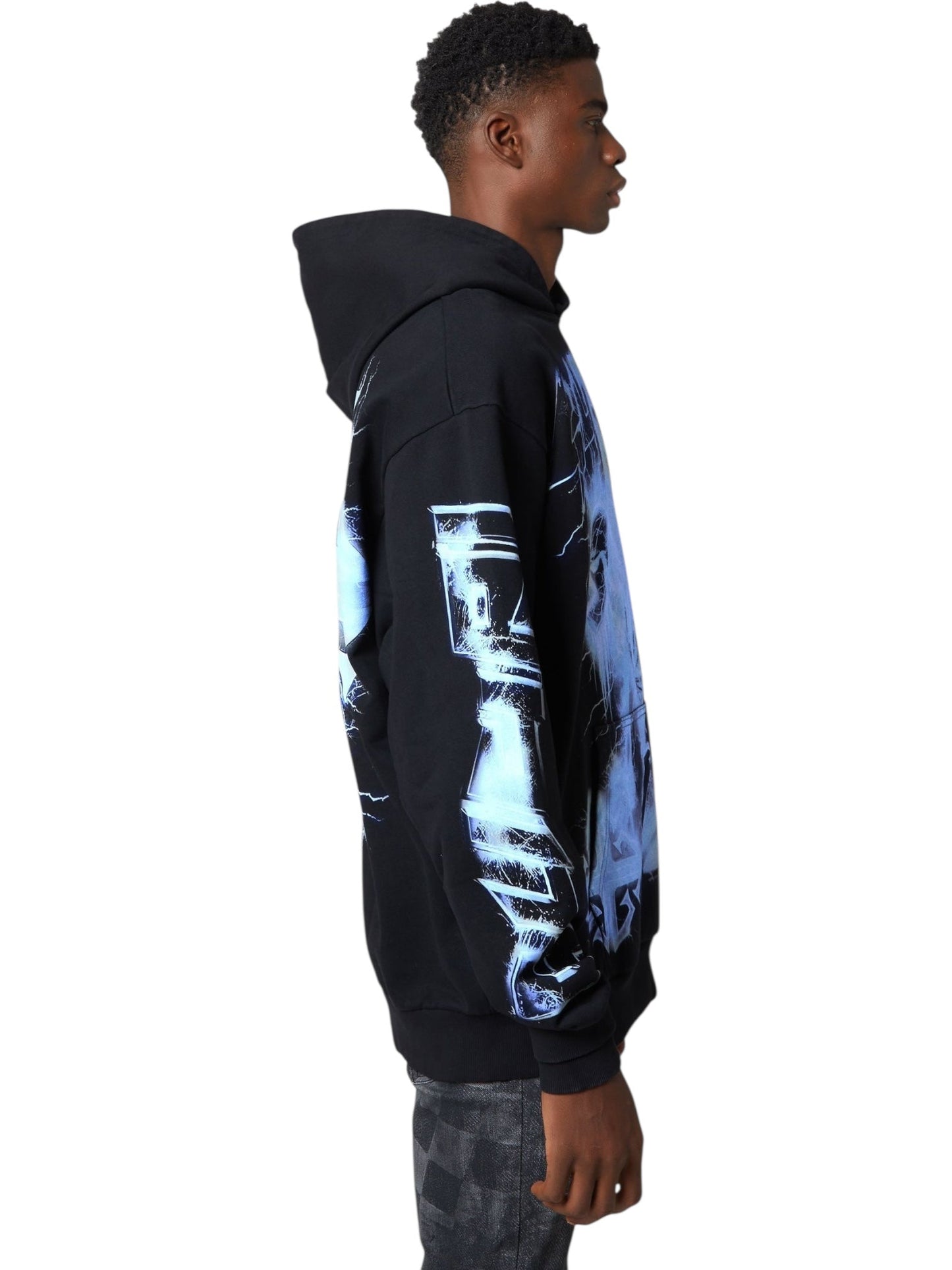 A young man stands in profile wearing the GUAPI Guapi Black Revived Hoodie V2 with blue graphic prints and dark patterned pants, showcasing modern streetwear style against a white background.