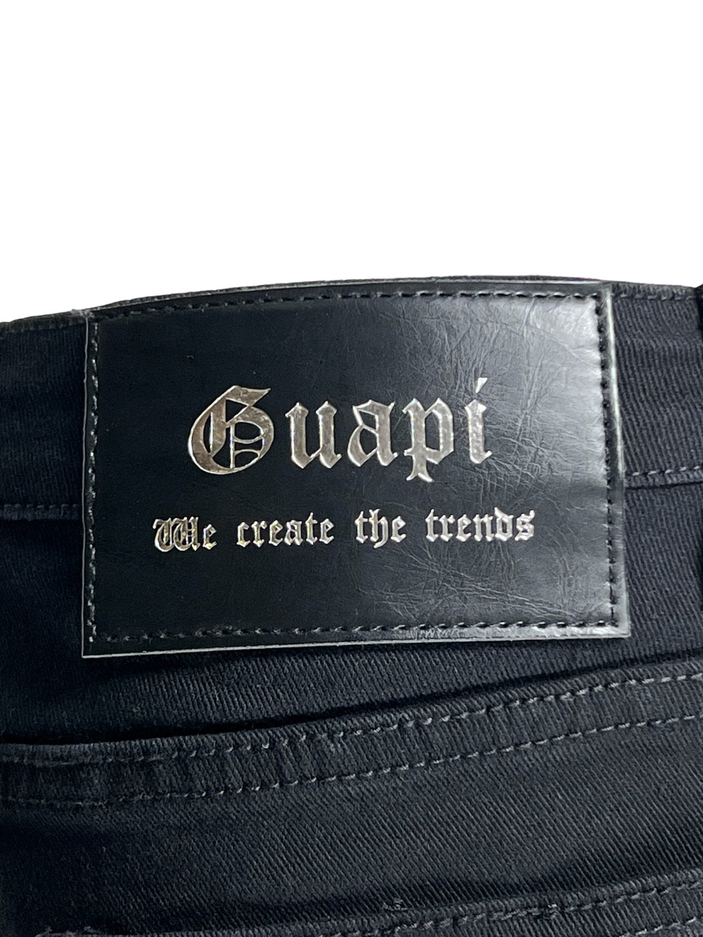 A close-up of a black leather label with the embossed text "GUAPI" and the slogan "we create the trends" on GUAPI Obsidian Black Blood Diamond Stacked Denim.