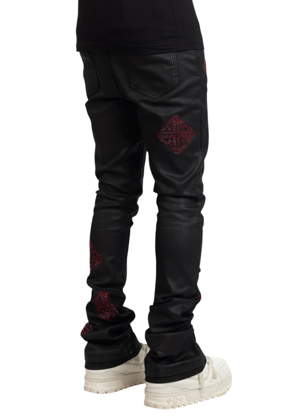 A person wears GUAPI's Obsidian Black Icon Embellished Denim V2 featuring red diamond patterns, paired with white sneakers, standing against a plain backdrop.