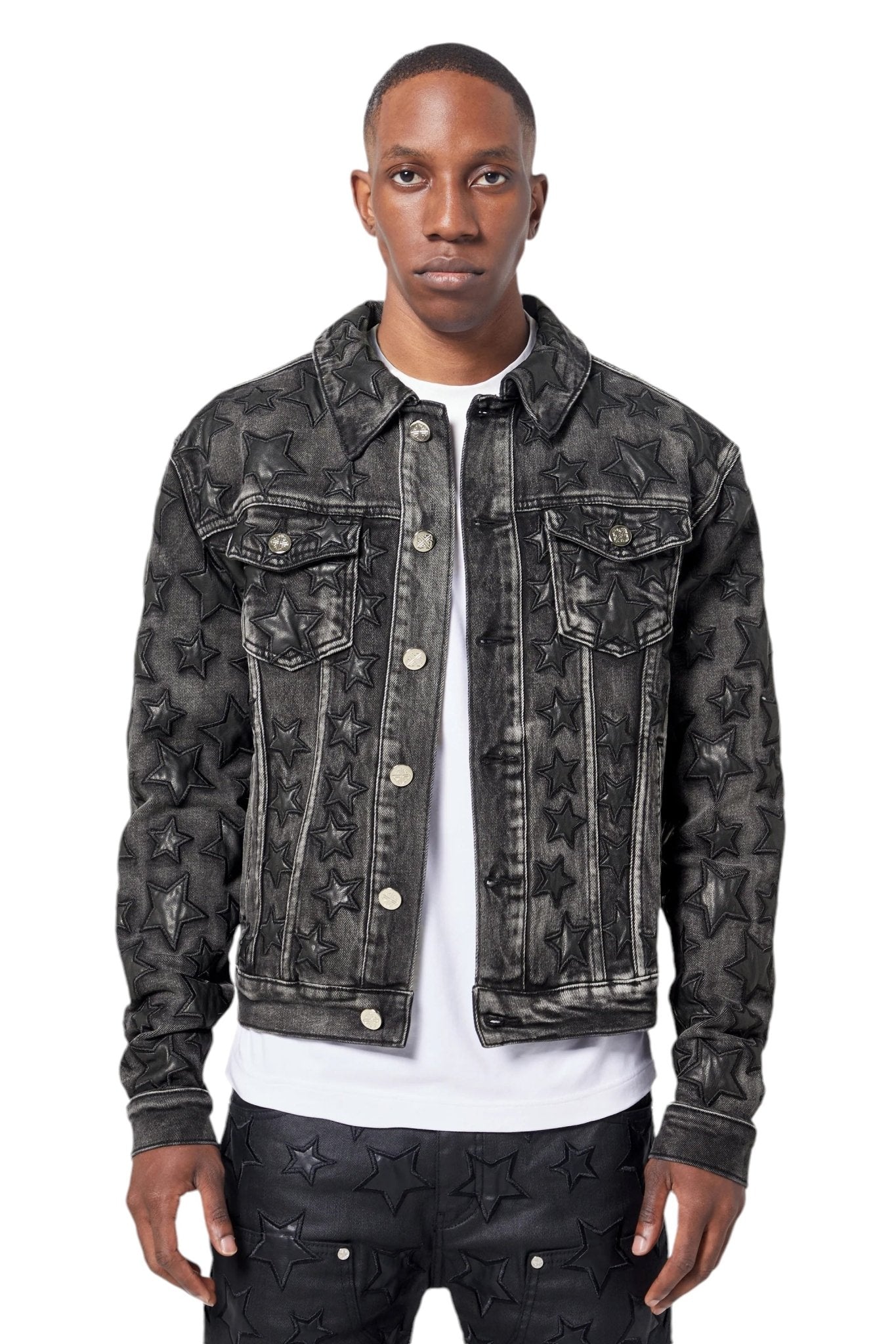 A person in a GUAPI Obsidian Black Stars Denim Jacket and matching pants, featuring leather star patches, stands against a plain white background, making a bold streetwear statement.