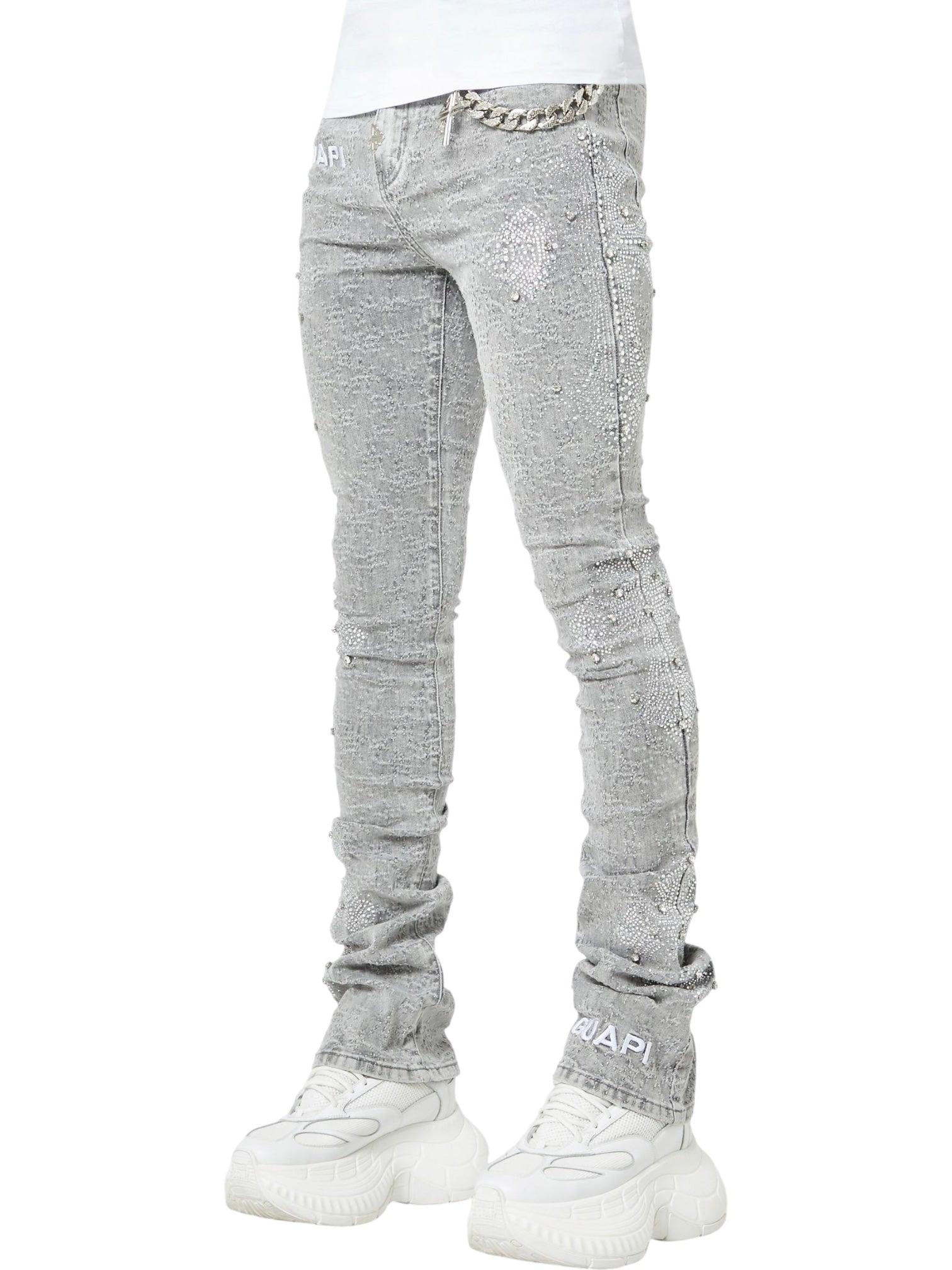 A person wears GUAPI Smoke Grey Distressed Cross Denim—light gray, textured skinny jeans with chain detail—paired with chunky white sneakers. Only the lower body is visible against a white background.