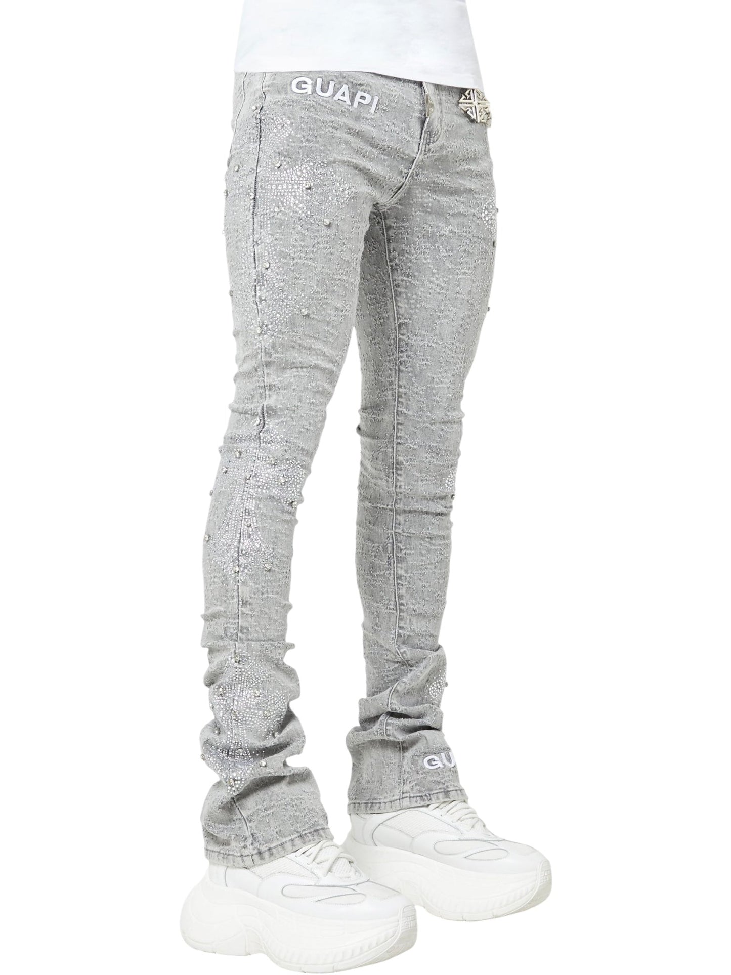 Model wearing GUAPI Smoke Grey Distressed Cross Denim stacked jeans with rhinestone cross details and chunky white sneakers, set against a plain background.