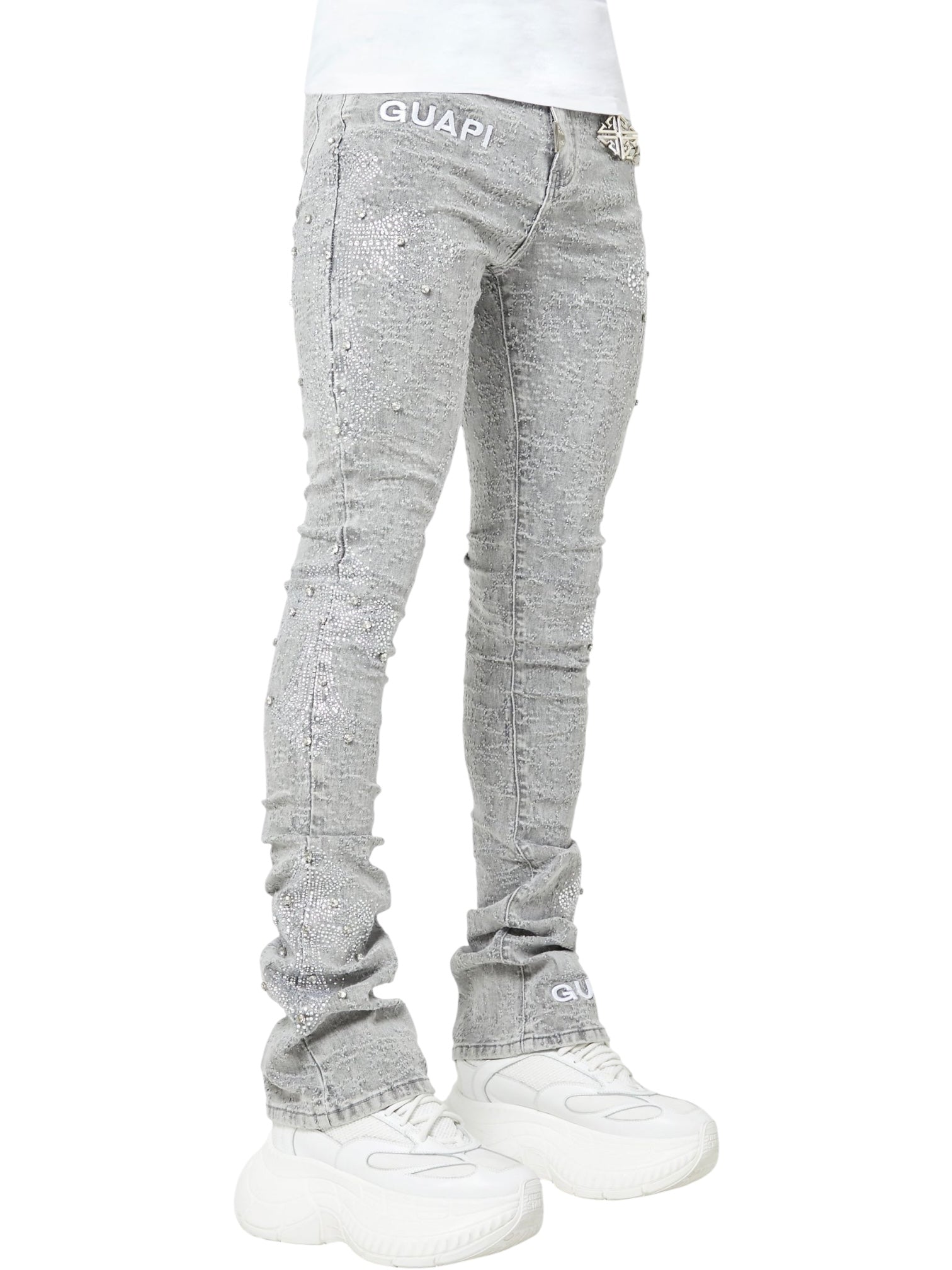 Model wearing GUAPI Smoke Grey Distressed Cross Denim stacked jeans with rhinestone cross details and chunky white sneakers, set against a plain background.