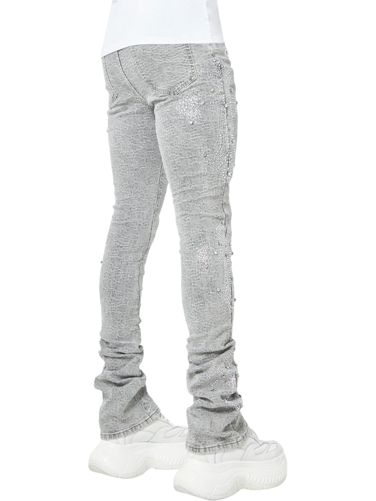 Someone wearing GUAPI's Guapi Smoke Grey Distressed Cross Denim, featuring rhinestone cross details, pairs the light gray jeans with chunky white sneakers; outfit is shown from the back and side.
