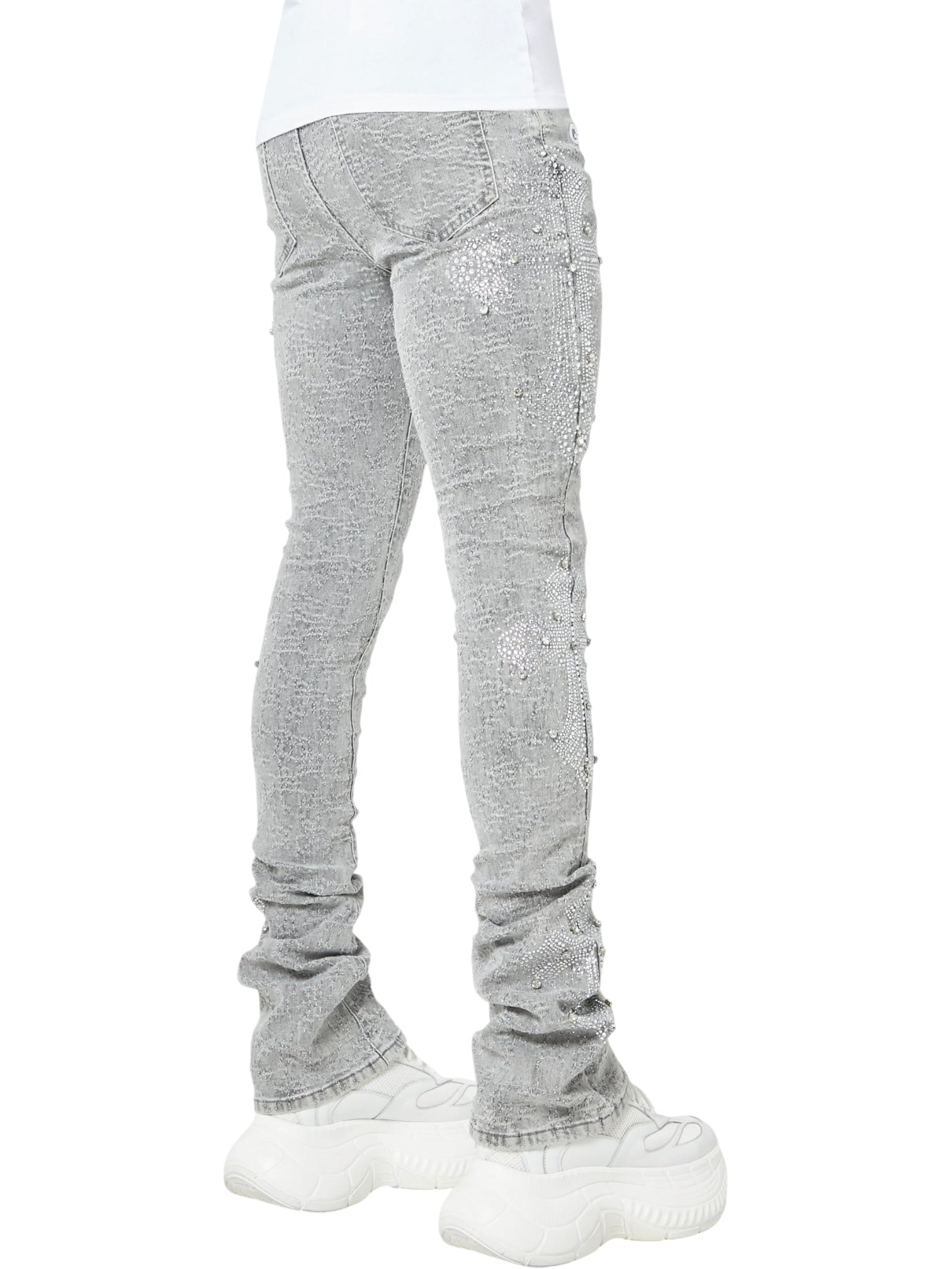 Someone wearing GUAPI's Guapi Smoke Grey Distressed Cross Denim, featuring rhinestone cross details, pairs the light gray jeans with chunky white sneakers; outfit is shown from the back and side.
